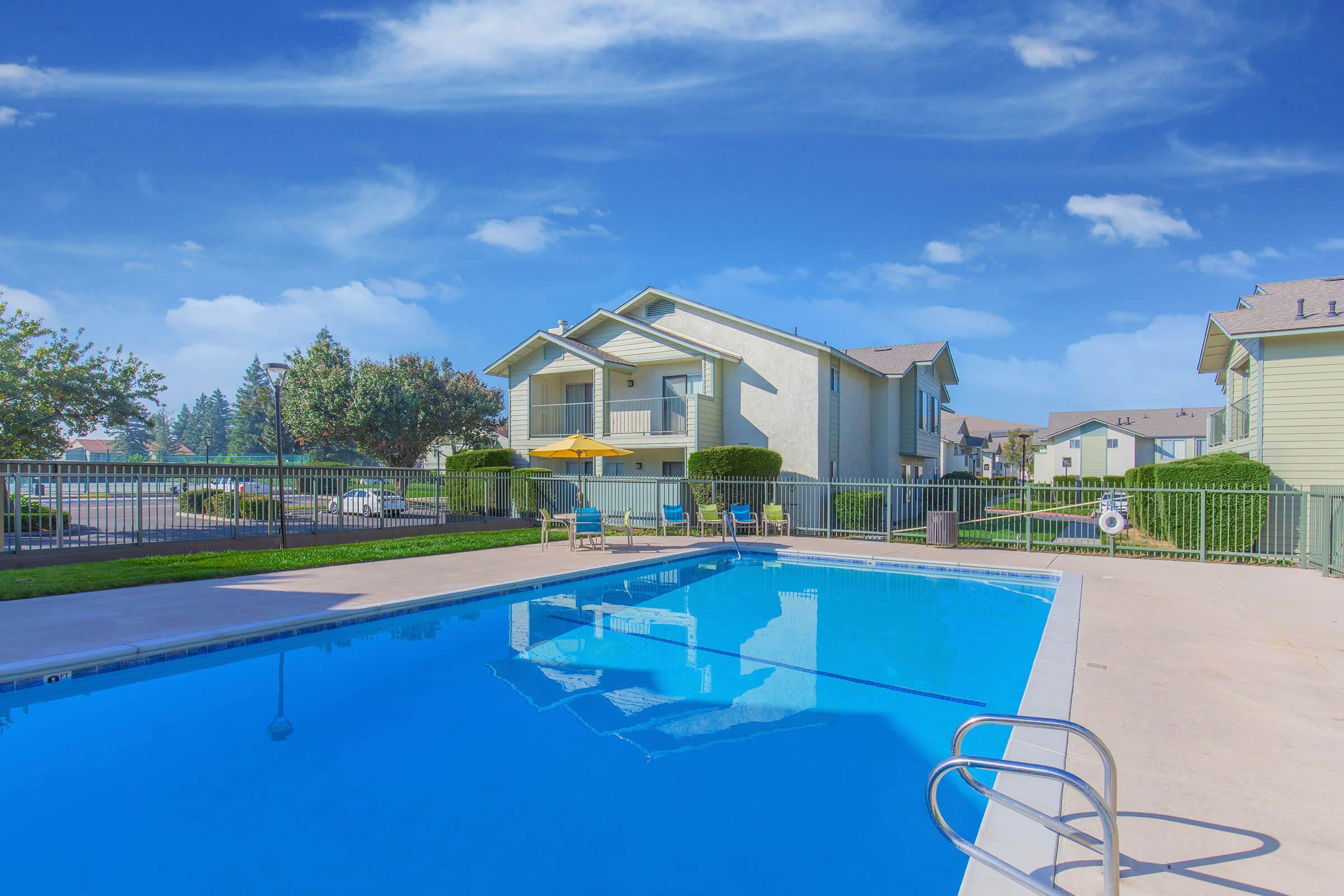 A serene outdoor swimming pool surrounded by a fence, featuring a diving board and a ladder. In the background, two-story apartment buildings with balconies and lush greenery are visible under a clear blue sky. Bright sunlight enhances the tranquil atmosphere.