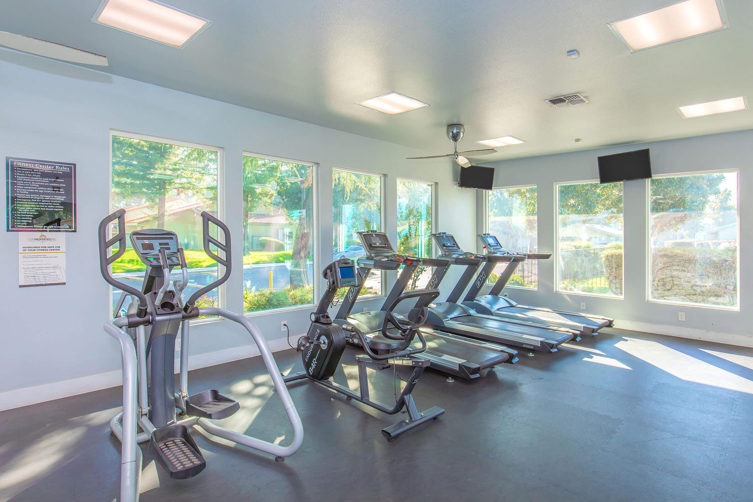 A bright, well-lit gym interior featuring several exercise machines, including treadmills and ellipticals, positioned near large windows that offer views of greenery outside. Wall-mounted TVs are visible, along with a fitness information poster on the wall. The space has a modern and inviting atmosphere.