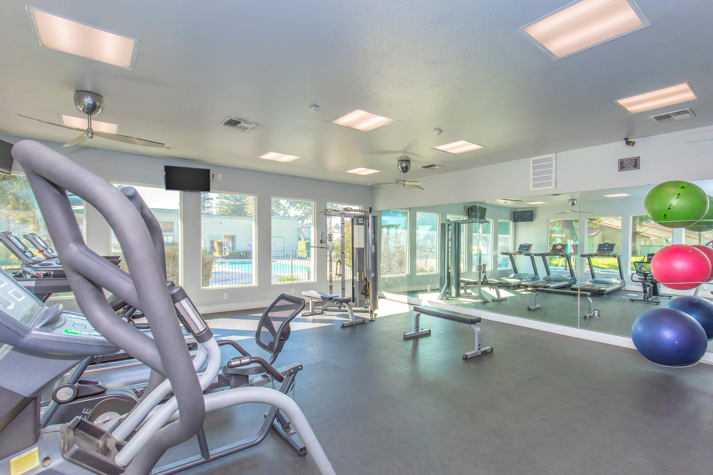 A modern gym interior featuring exercise equipment such as treadmills and elliptical machines, along with colorful stability balls. Large windows provide natural light, and the space is designed for fitness activities with a mirrored wall enhancing the sense of openness.