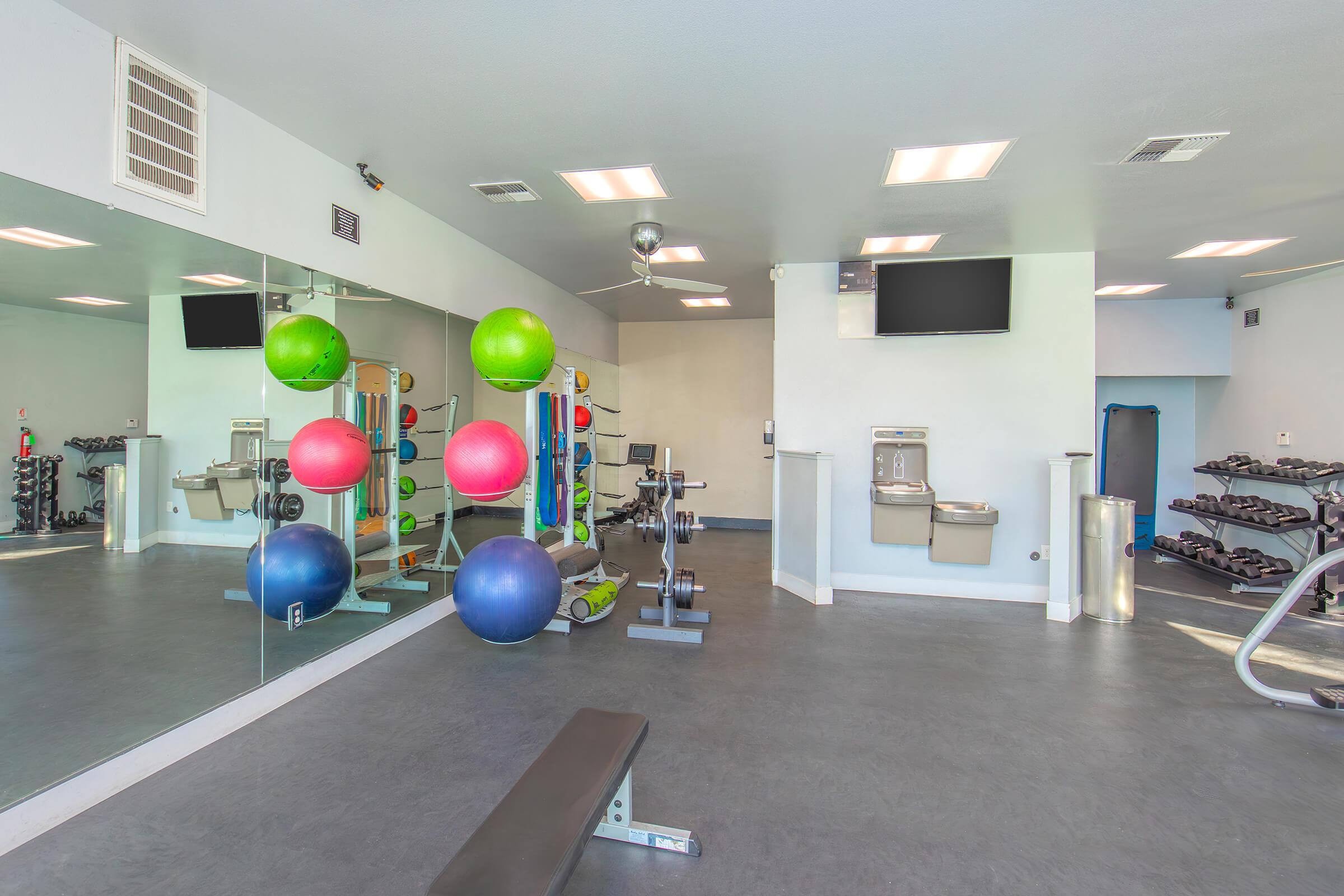 A well-lit gym interior featuring exercise equipment, including dumbbells, a weight bench, and various gym balls in vibrant colors. Mirrors on one wall reflect the space, and there are two televisions mounted on the wall. A water fountain is visible, enhancing the overall fitness environment.