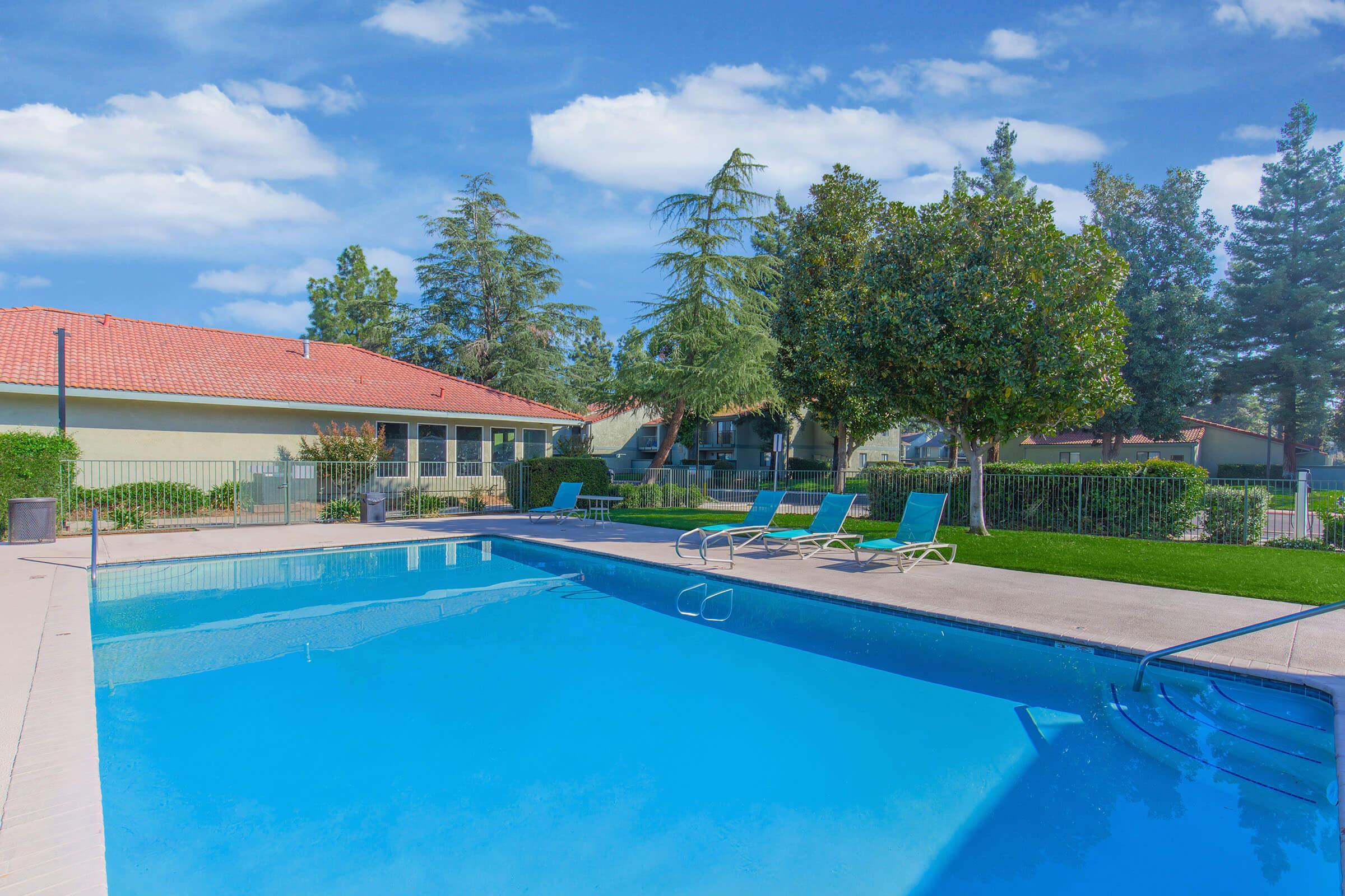 A bright and inviting outdoor swimming pool surrounded by lounge chairs and lush greenery. The pool features a clear blue water surface, with a well-maintained patio area and nearby residential buildings under a blue sky with fluffy clouds.
