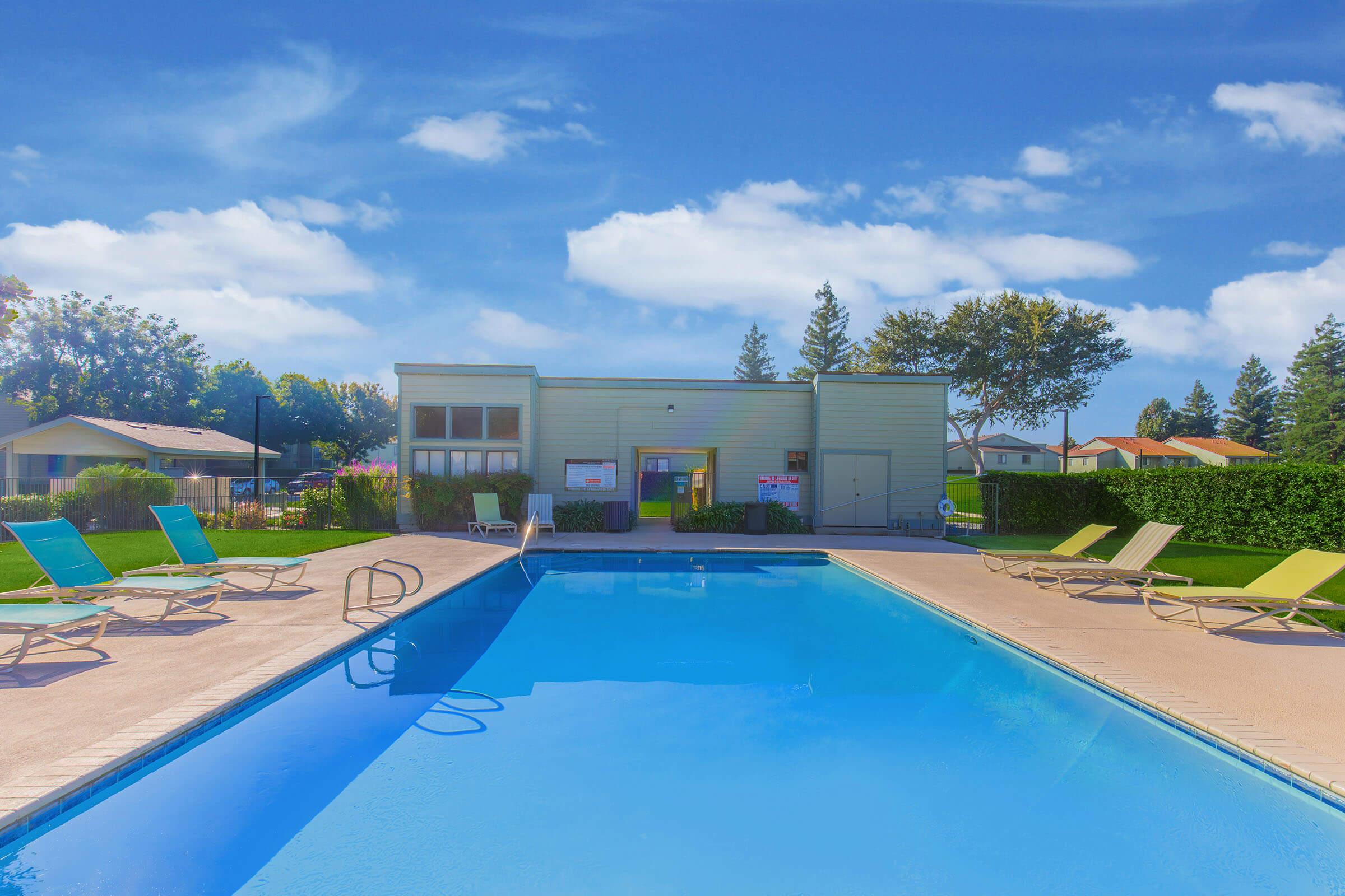 A clear blue swimming pool surrounded by lounge chairs, with a well-maintained lawn and small trees in the background. In the distance, a single-story building with windows and a poolside area can be seen against a bright blue sky with a few clouds.