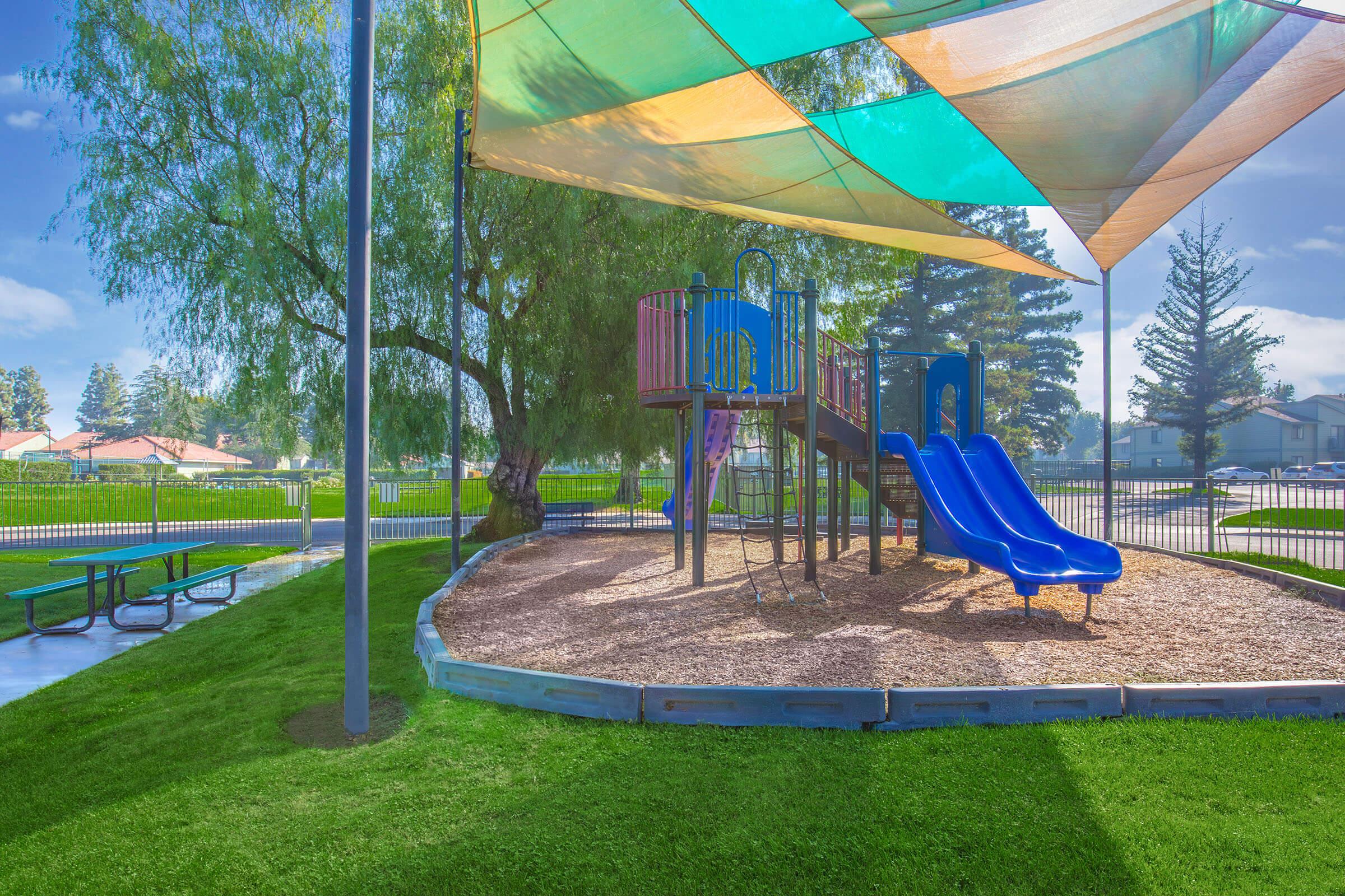 A colorful playground with a slide and climbing structure, shaded by bright canopies, surrounded by lush green grass and a gravel area. Nearby, there are picnic tables under a shaded area, with trees and a fence in the background. The scene is bright and inviting, perfect for children's outdoor play.