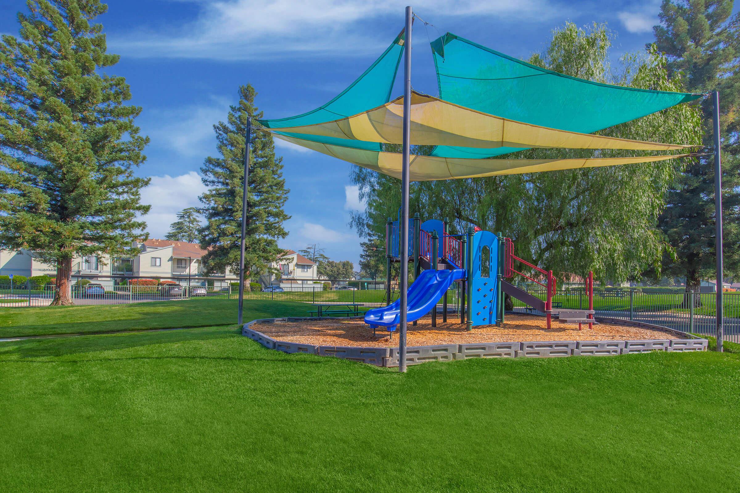 A colorful playground with slides and climbing structures, shaded by large canopies. The area is surrounded by green grass and trees, with houses visible in the background under a clear blue sky. Ideal for children to play and enjoy outdoor activities.