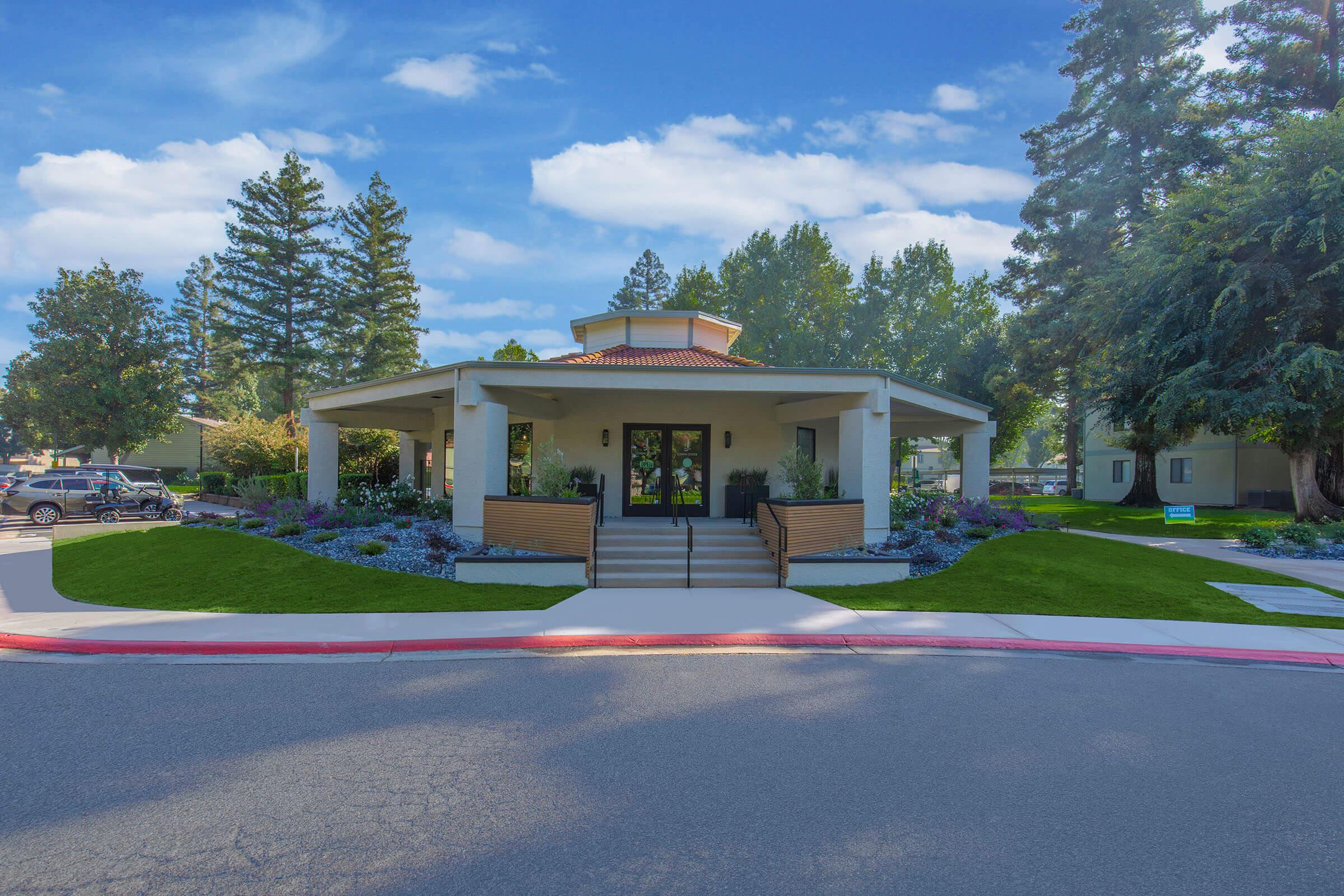 A modern building with a round roof and entrance surrounded by landscaped gardens and colorful flowers. The structure features a porch with steps leading up to the entrance, framed by neatly trimmed bushes. Tall trees are visible in the background, creating a serene atmosphere.