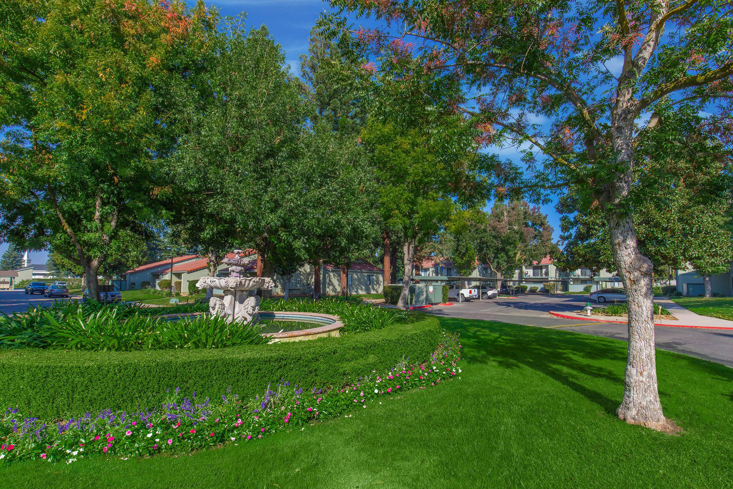 A landscaped area featuring a circular flower bed with colorful flowers, surrounded by lush green grass and trees. In the background, a residential complex can be seen, with parked cars along a paved road. The sky is clear and blue, creating a serene outdoor environment.