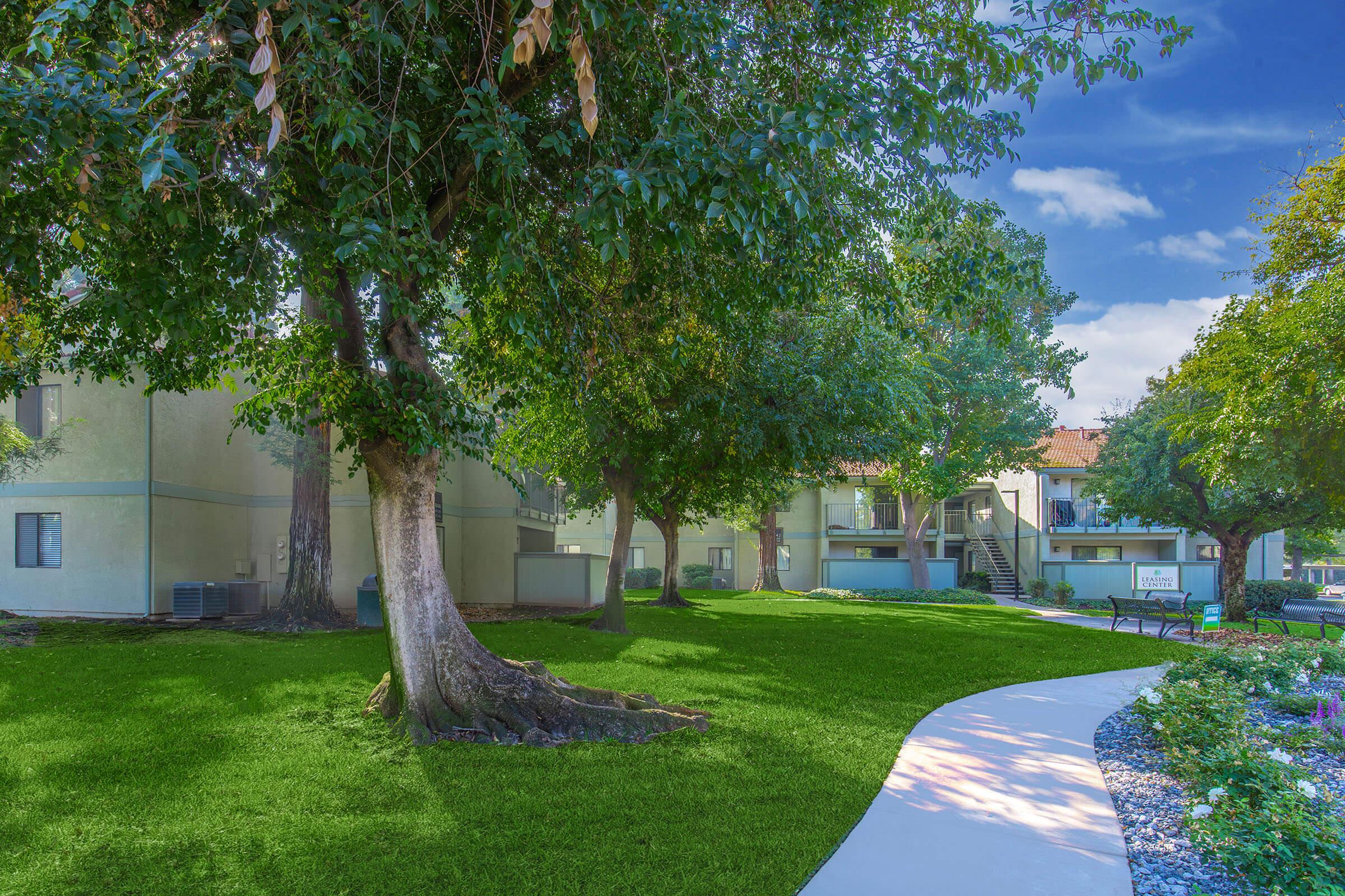 A well-maintained apartment complex surrounded by lush greenery, featuring large trees and a manicured lawn. A winding path leads through the area, with benches for residents to relax. The sky is bright and blue, enhancing the inviting atmosphere of the community.