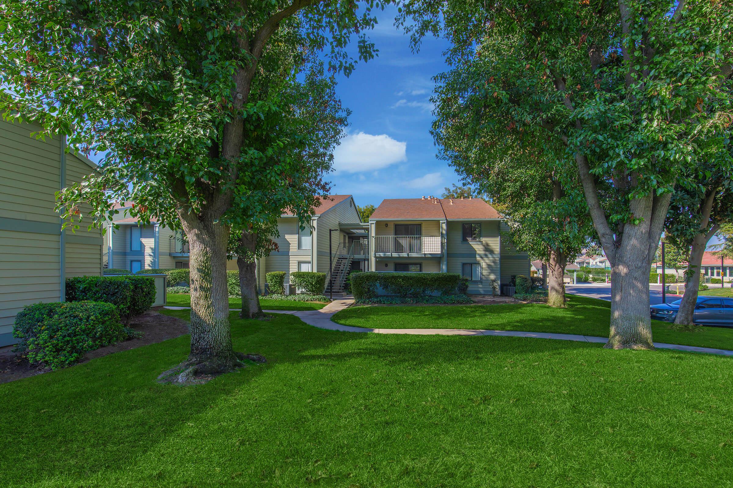 A well-maintained apartment complex surrounded by lush green grass and trees. The building features a staircase leading to upper-level units, with neatly trimmed hedges and a clear blue sky in the background. Pathways meander through the landscape, creating an inviting atmosphere.