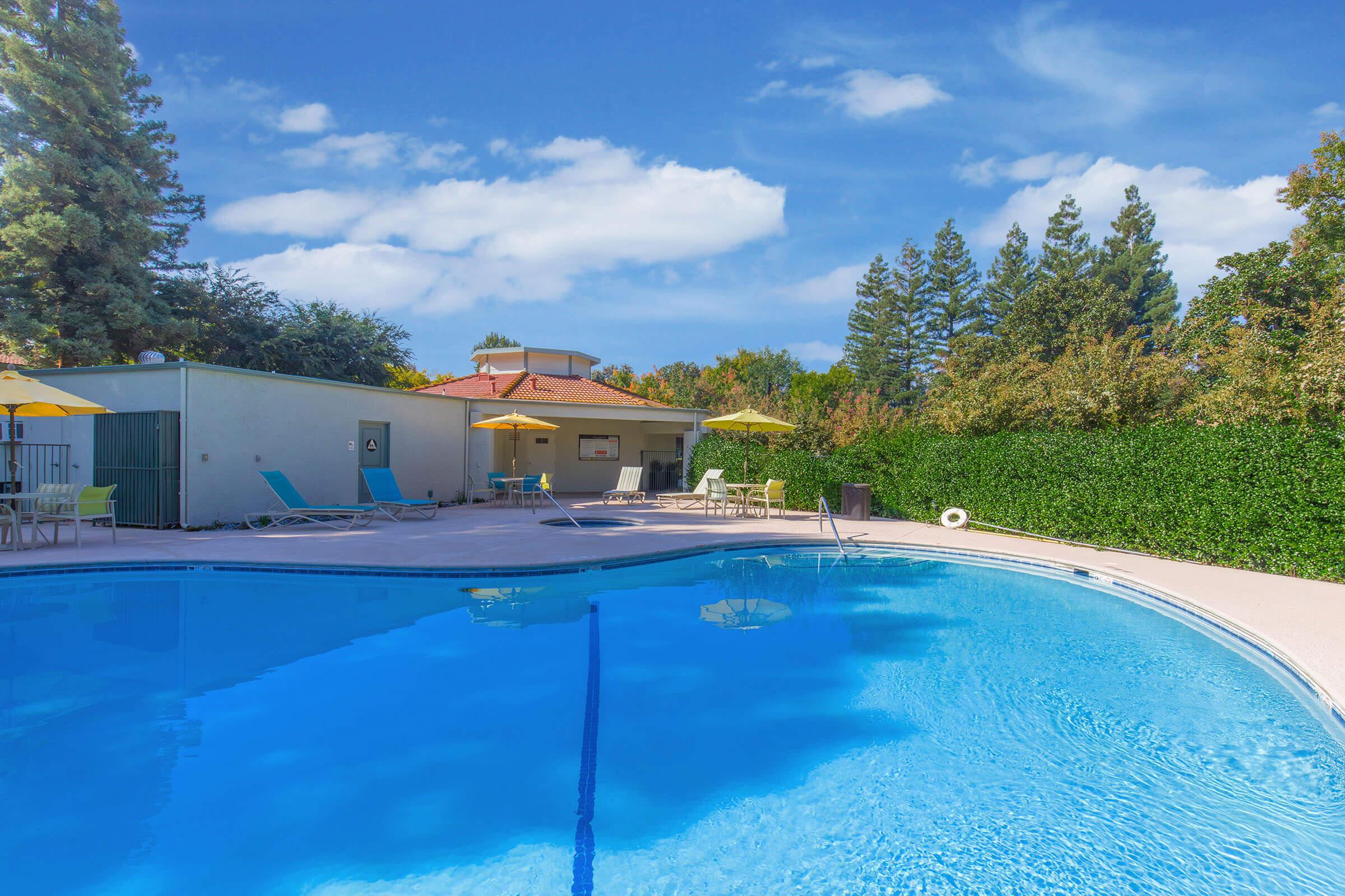 A clear blue swimming pool surrounded by lounge chairs and yellow umbrellas. In the background, there is a building with a red roof, bordered by green hedges and tall trees, under a bright blue sky with fluffy white clouds. The scene is inviting and perfect for relaxation.