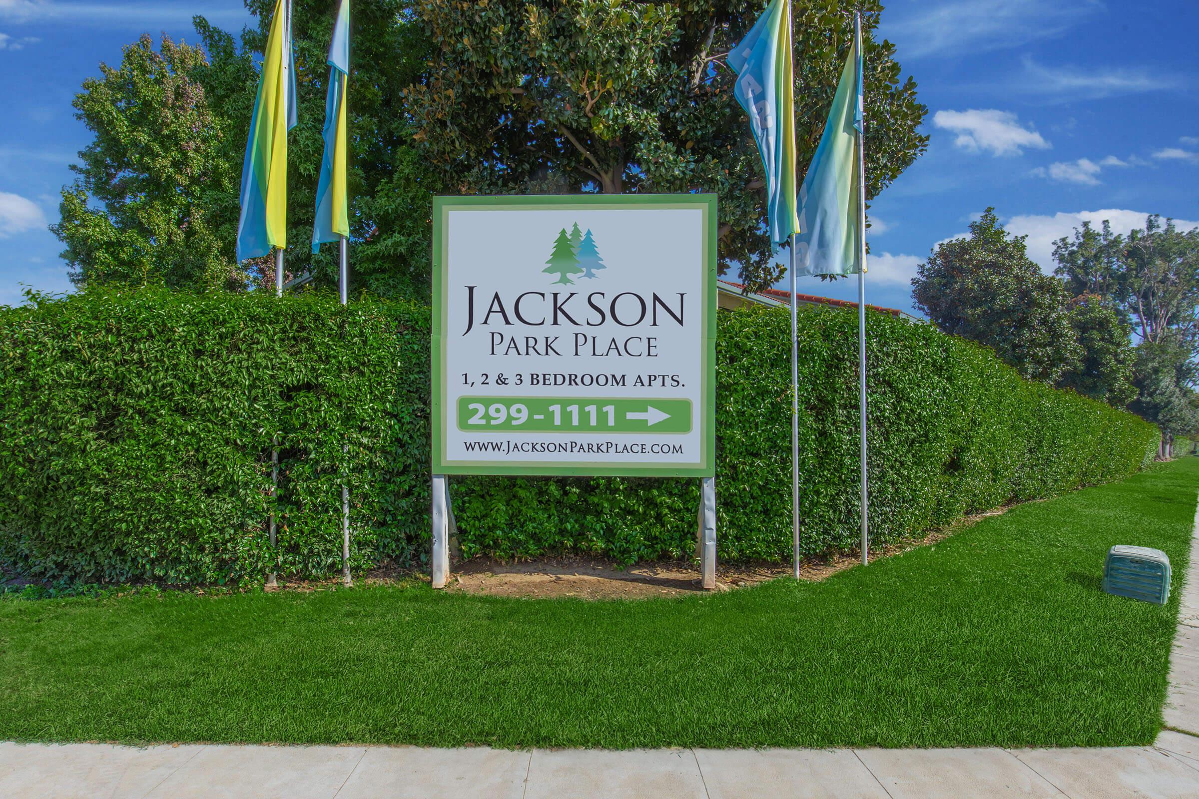 Sign for Jackson Park Place, promoting 1, 2, and 3-bedroom apartments. Features a green backdrop with trees and flags, located in a landscaped area. Phone number displayed as 299-1111, along with the website www.jacksonparkplace.com.