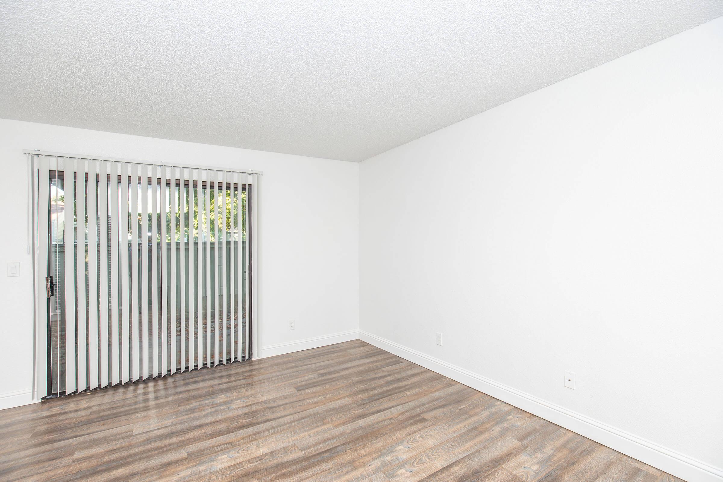 A spacious room with light-colored walls and wood laminate flooring. A large sliding glass door with vertical blinds leads to an outdoor space. The room is empty, creating a bright and open atmosphere, suitable for various uses.
