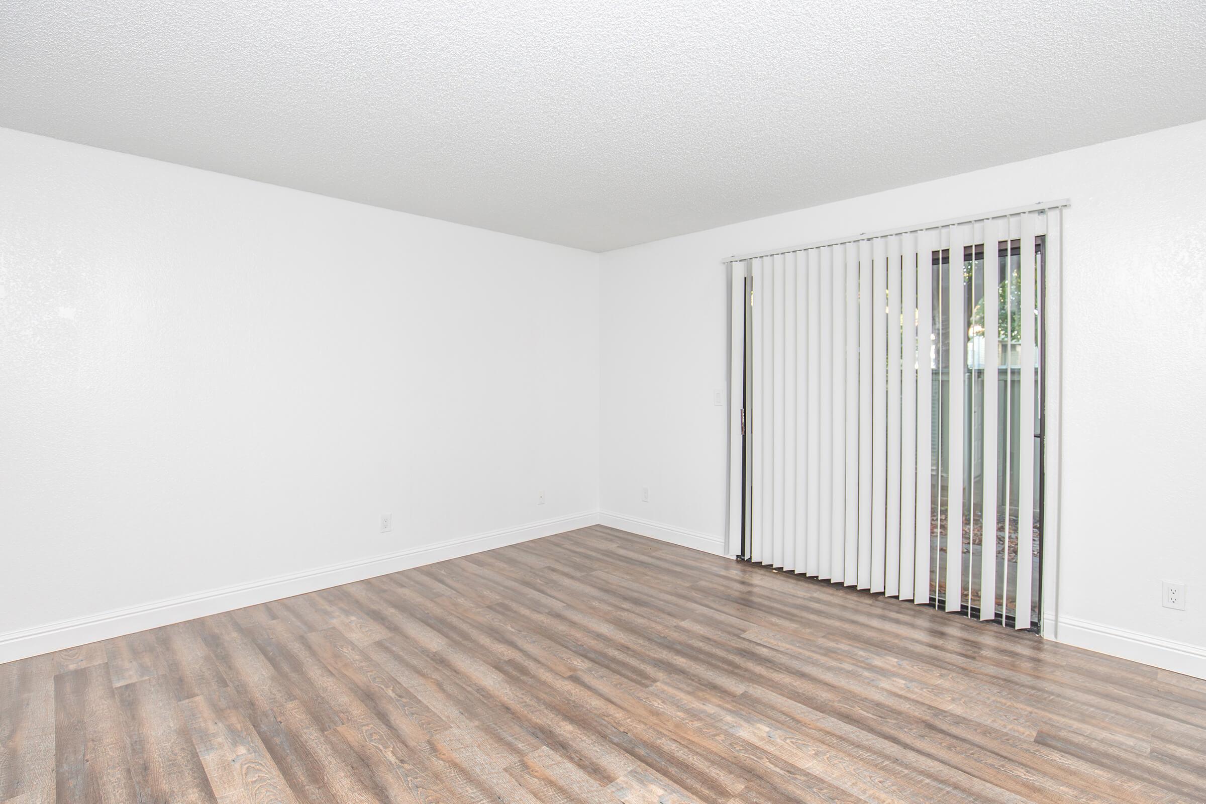 Empty room with white walls and a wooden floor. There is a large window with vertical blinds allowing natural light, and a sliding glass door that opens to an outdoor space. The room appears spacious and unadorned, ideal for personalization or decoration.