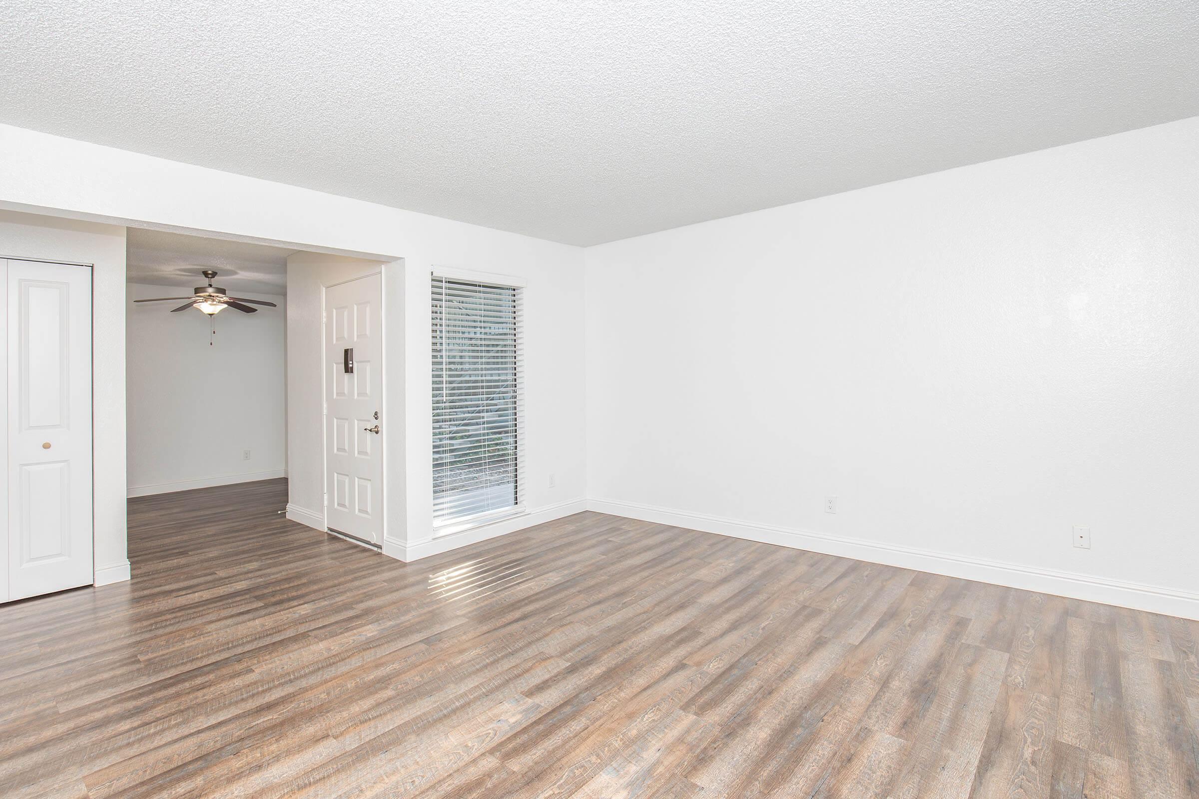 A spacious, empty living room with light-colored walls and wood-like flooring. It features a ceiling fan, a large window with blinds, and an open doorway leading to another room. The area is well-lit, creating a bright and airy atmosphere.