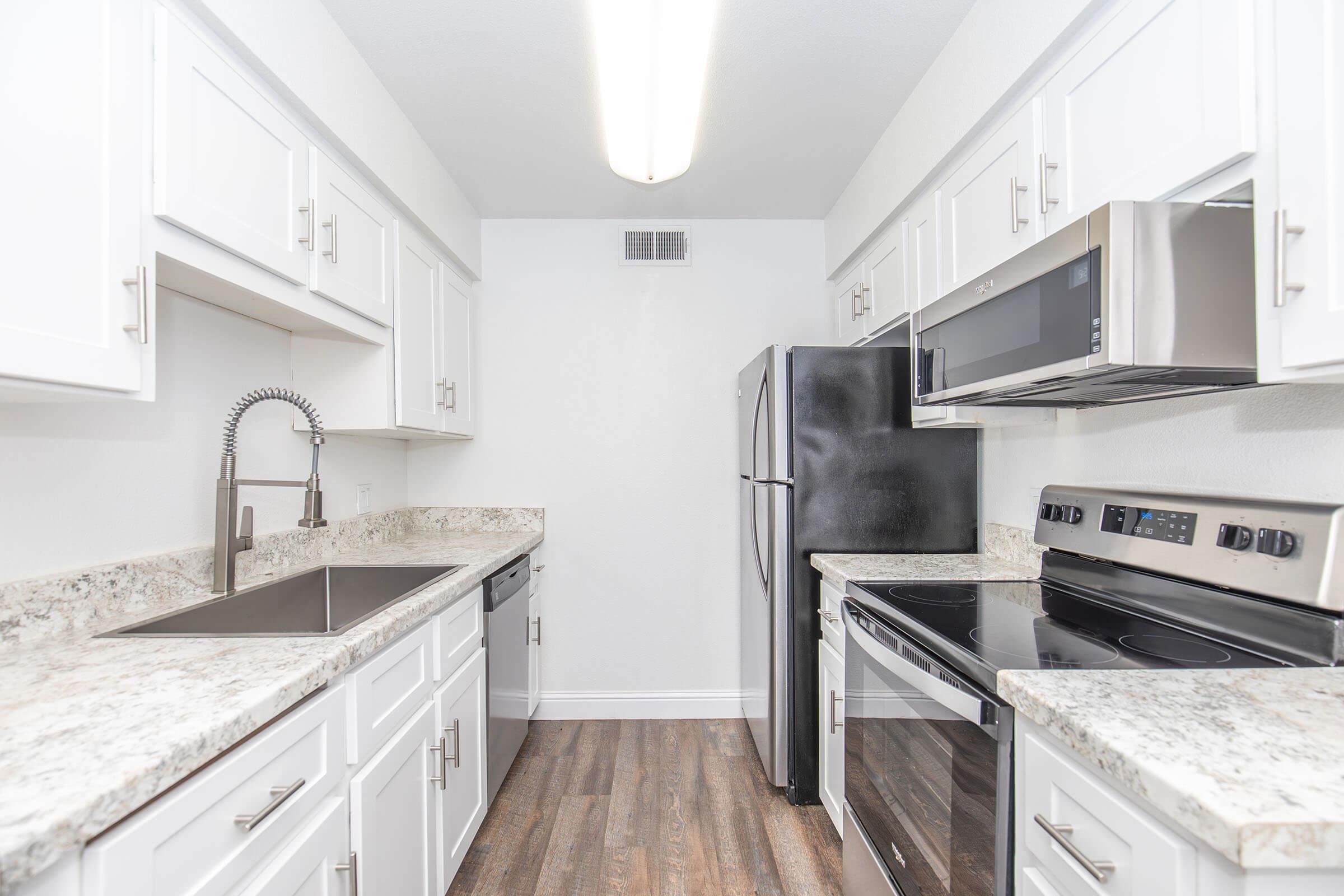 Modern kitchen featuring white cabinetry, granite countertops, stainless steel appliances including an oven, microwave, and refrigerator, along with a deep sink and a spacious layout, all set against a light-colored wall.