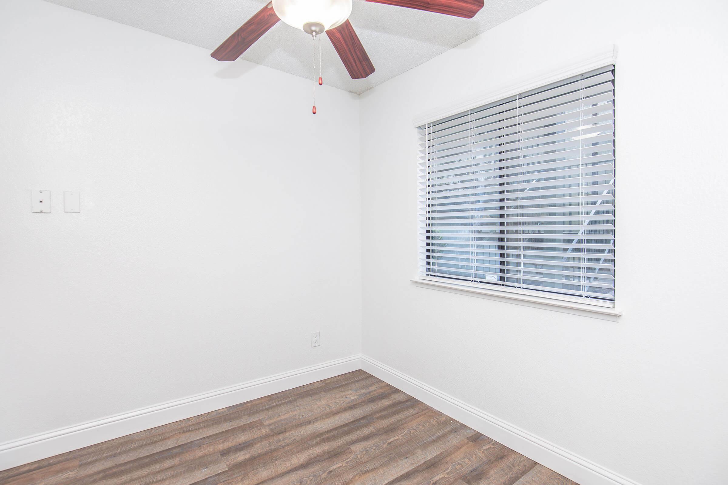 A bright, empty room featuring light-colored walls and a wooden floor. A ceiling fan with a light fixture hangs from the ceiling, and a window with horizontal blinds allows natural light to enter. The space is uncluttered, creating a clean and open atmosphere.