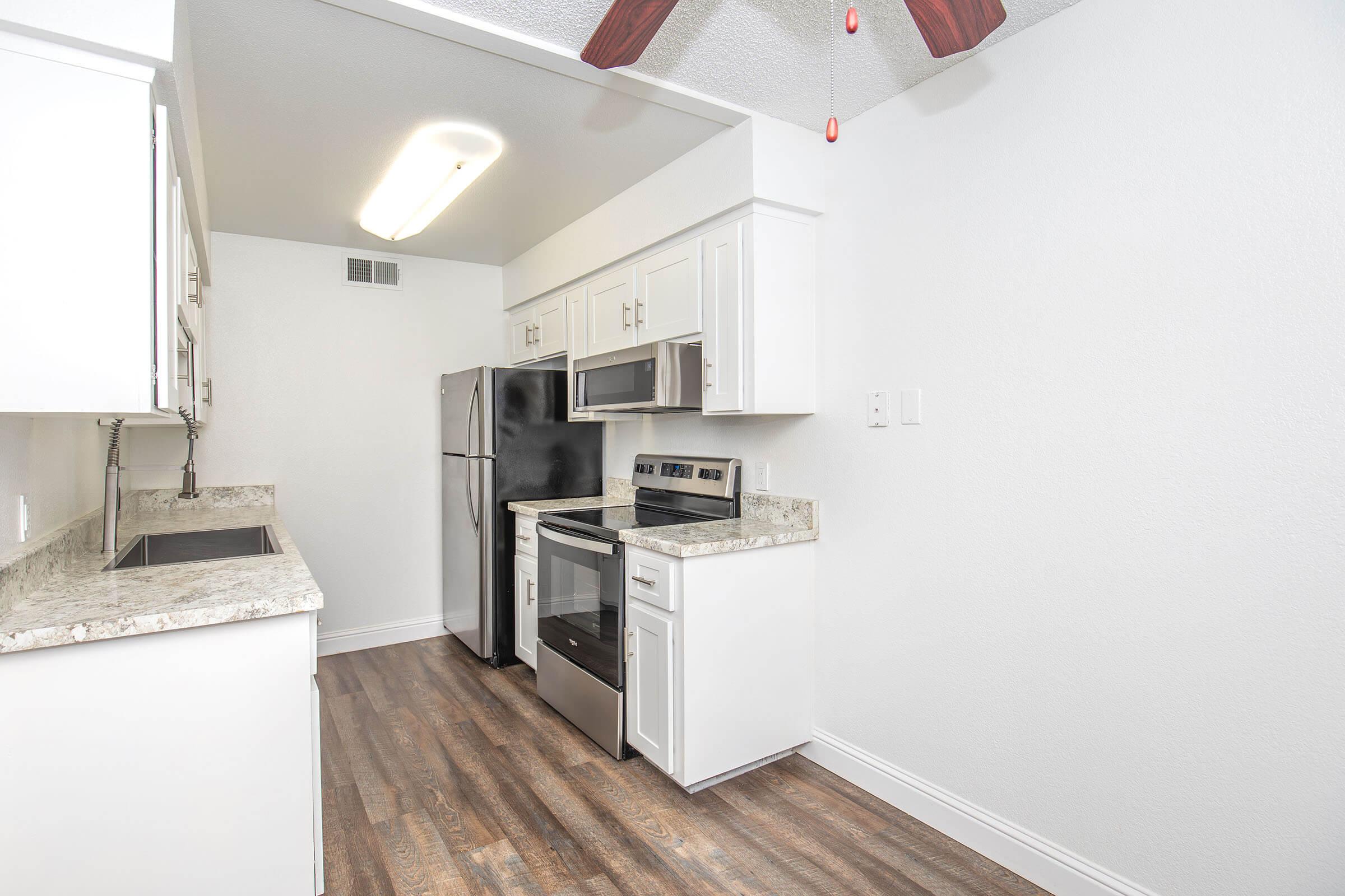 A modern kitchen featuring white cabinetry, a stainless steel refrigerator, microwave, and oven, with a granite countertop. The space has a light-colored wall and a ceiling fan above, with laminate flooring that adds a warm touch.