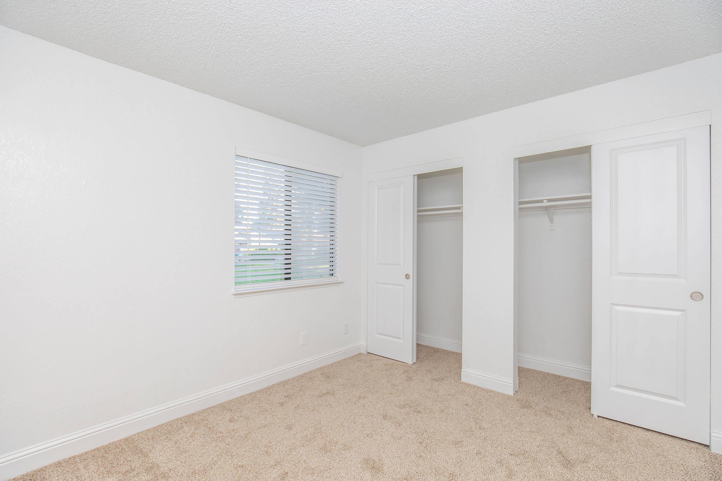 A well-lit, empty bedroom featuring light-colored walls and beige carpet. The room has a window with blinds on one side and two closet doors on the opposite side, providing ample storage space.