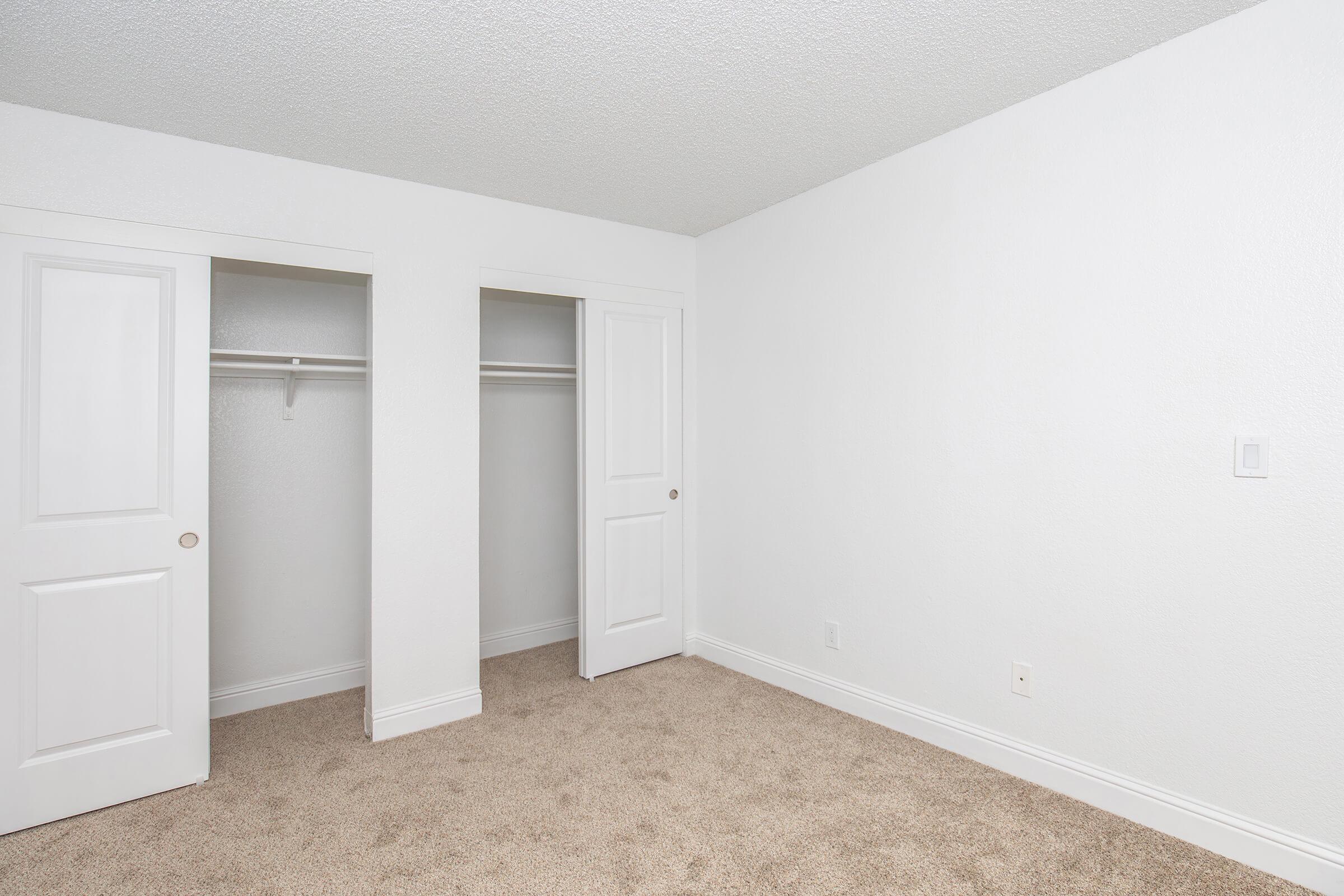 A spacious, empty room with beige carpet and white walls. It features two closed closet doors on the left side and one more door on the right side, all with simple white trim. The room has a clean, minimalist design, with no furniture or decorations present.