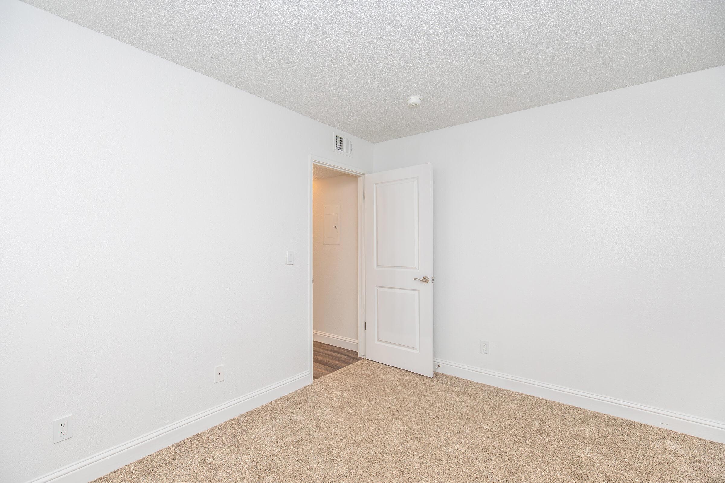 A well-lit, empty room with white walls and a light-colored carpet. A closed door is visible on the left, leading to another space. The room is sparsely decorated, offering a clean and minimalistic look, perfect for various uses.