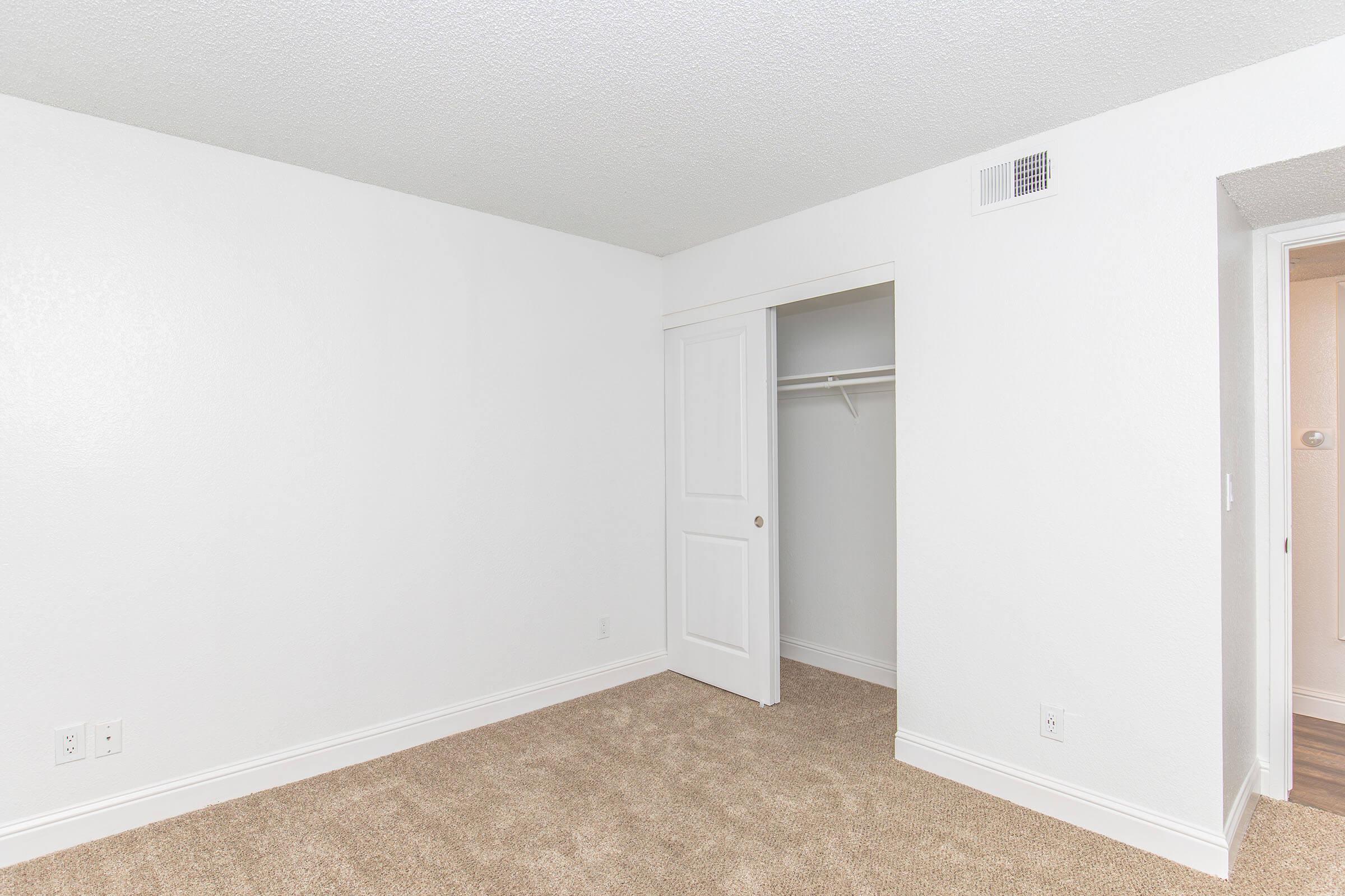 A bright, empty room with white walls and carpeted flooring. There is a closed door leading to a closet on one side, and a doorway that appears to lead to another area, possibly a bathroom. The overall ambiance is clean and modern.