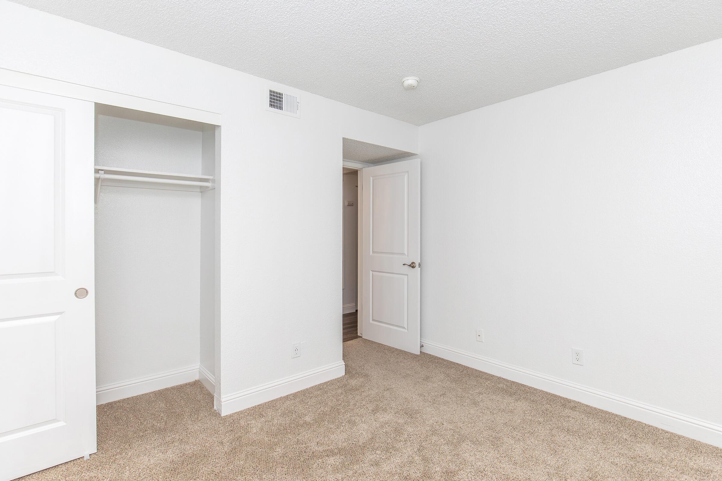 A small, empty room featuring beige carpet and white walls. There is a closet with a white door on the left, and a second door leading to another space on the right. The room is well-lit with a ceiling light, creating a clean and neutral atmosphere.