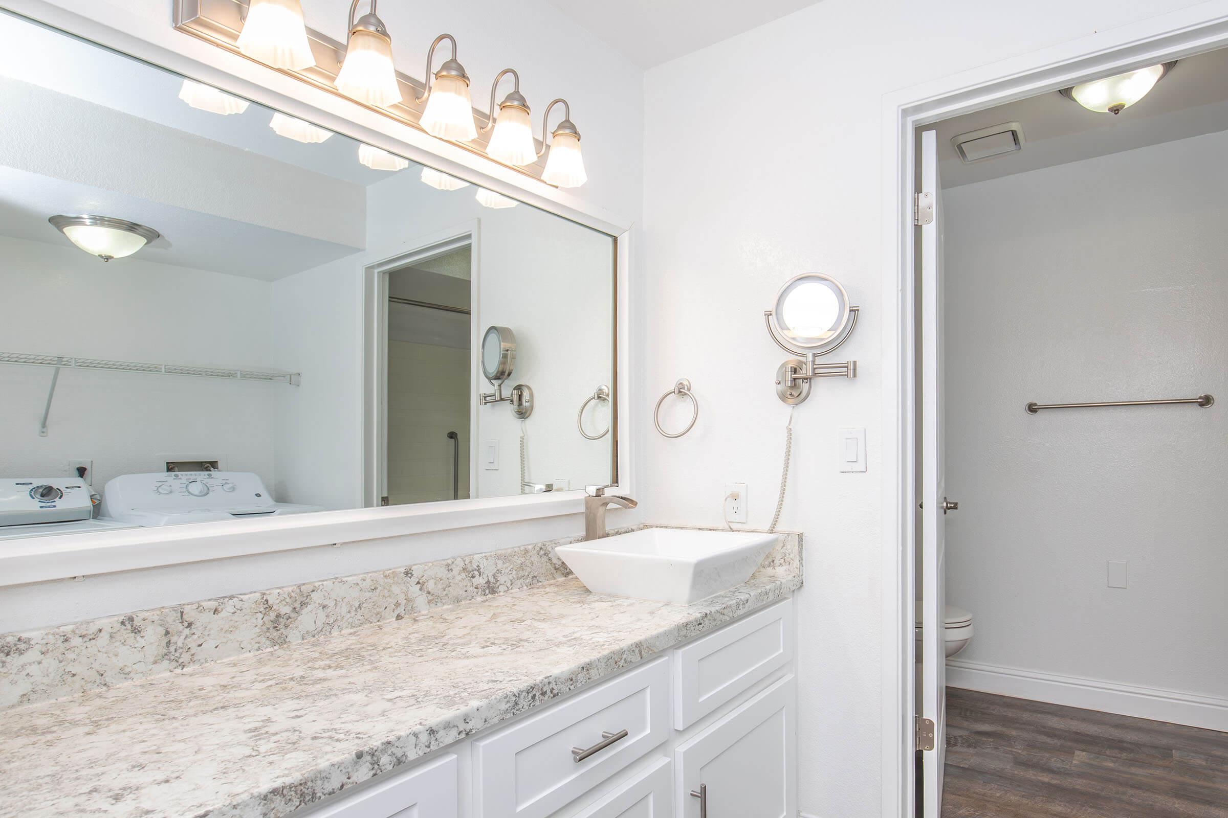 A modern bathroom featuring a granite countertop with a rectangular sink, a large mirror with five light fixtures above, and a wall-mounted mirror. To the left, there is a laundry area with a washing machine. The room is painted white, creating a clean, bright atmosphere, and includes a door leading to a small toilet area.