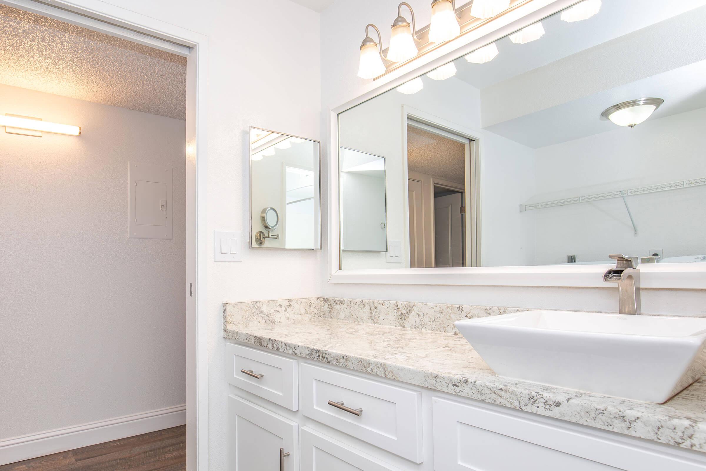 Modern bathroom featuring a white vanity with a granite countertop, a stylish vessel sink, and a large mirror above. The space is brightly lit with overhead lights and has light-colored walls, giving it a clean and contemporary look. A door leads to another area, adding to the room's spacious feel.