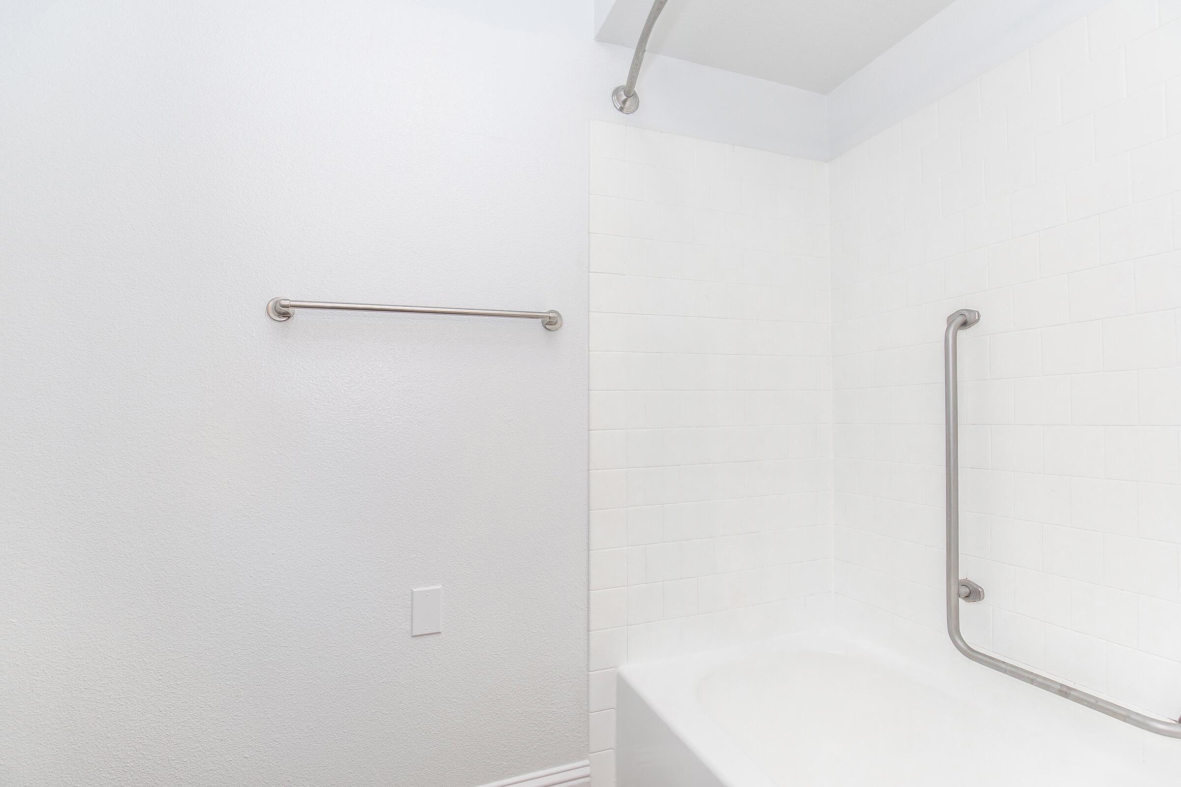 A clean, modern bathroom featuring a white tiled shower area with a tub. There are grab bars installed on the walls for safety, and a towel rod is mounted beside the tub. The walls are painted in a light color, creating a bright and airy atmosphere.