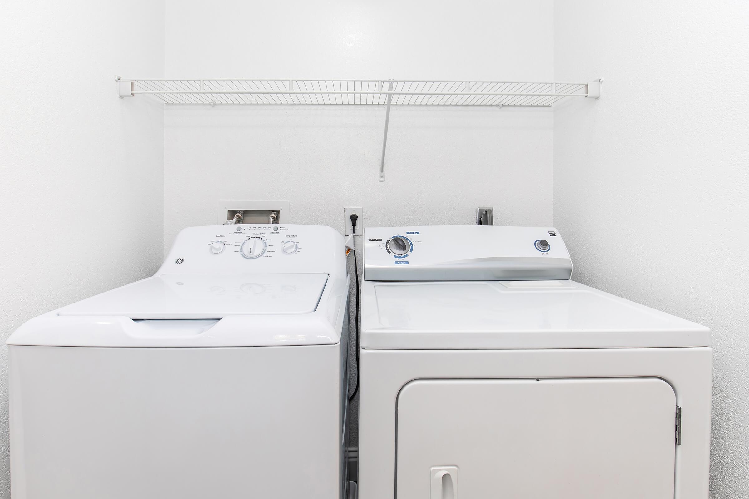 A bright laundry room featuring a white washing machine and a white dryer side by side. Above them is a wire shelf mounted on the wall, and the walls are painted white, creating a clean and modern look.