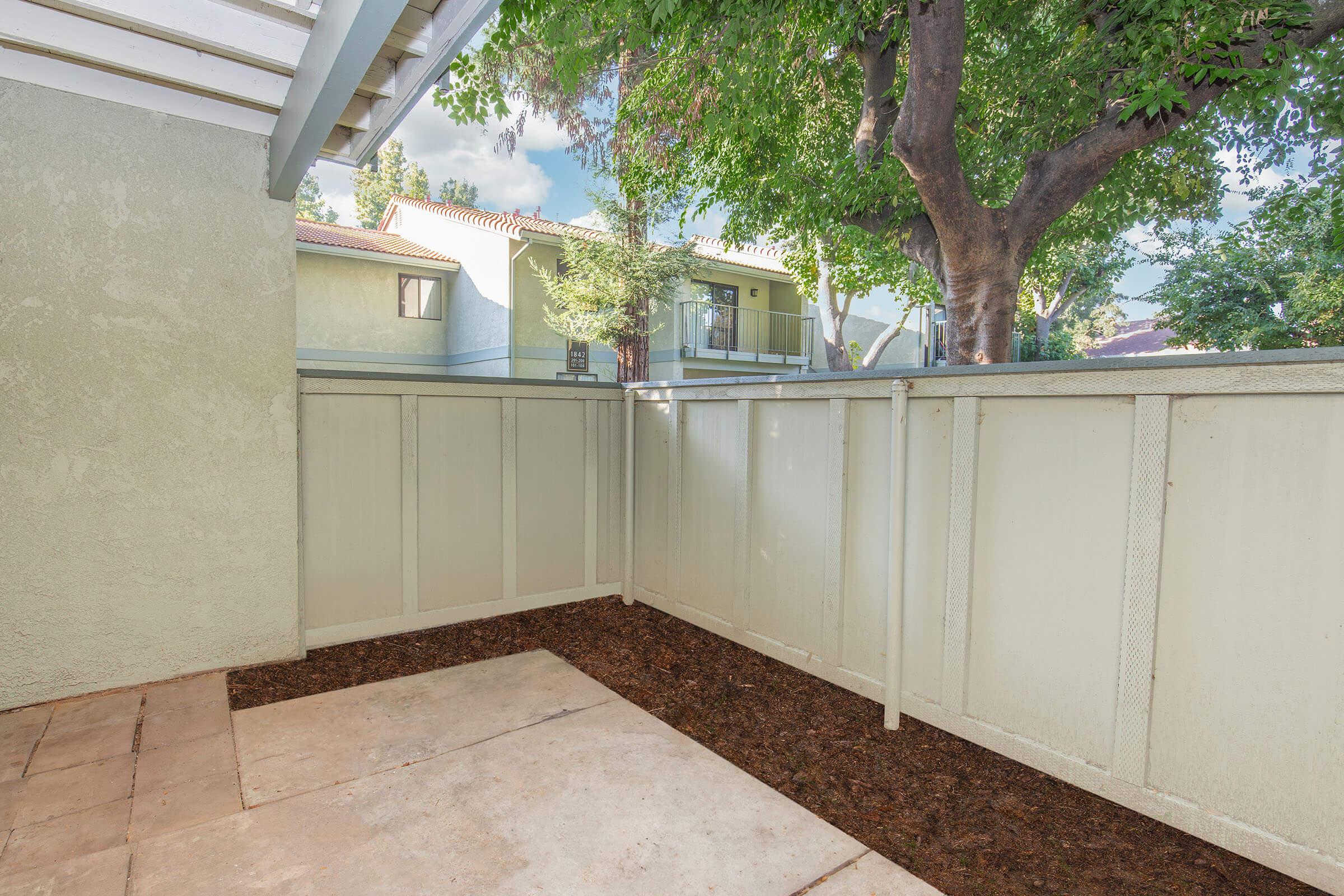 A small, enclosed outdoor patio area with a gravel floor, surrounded by light-colored wooden fencing. Green trees and shrubs are visible in the background, along with the exterior of nearby residential buildings. The scene is well-lit, suggesting a pleasant daytime atmosphere.