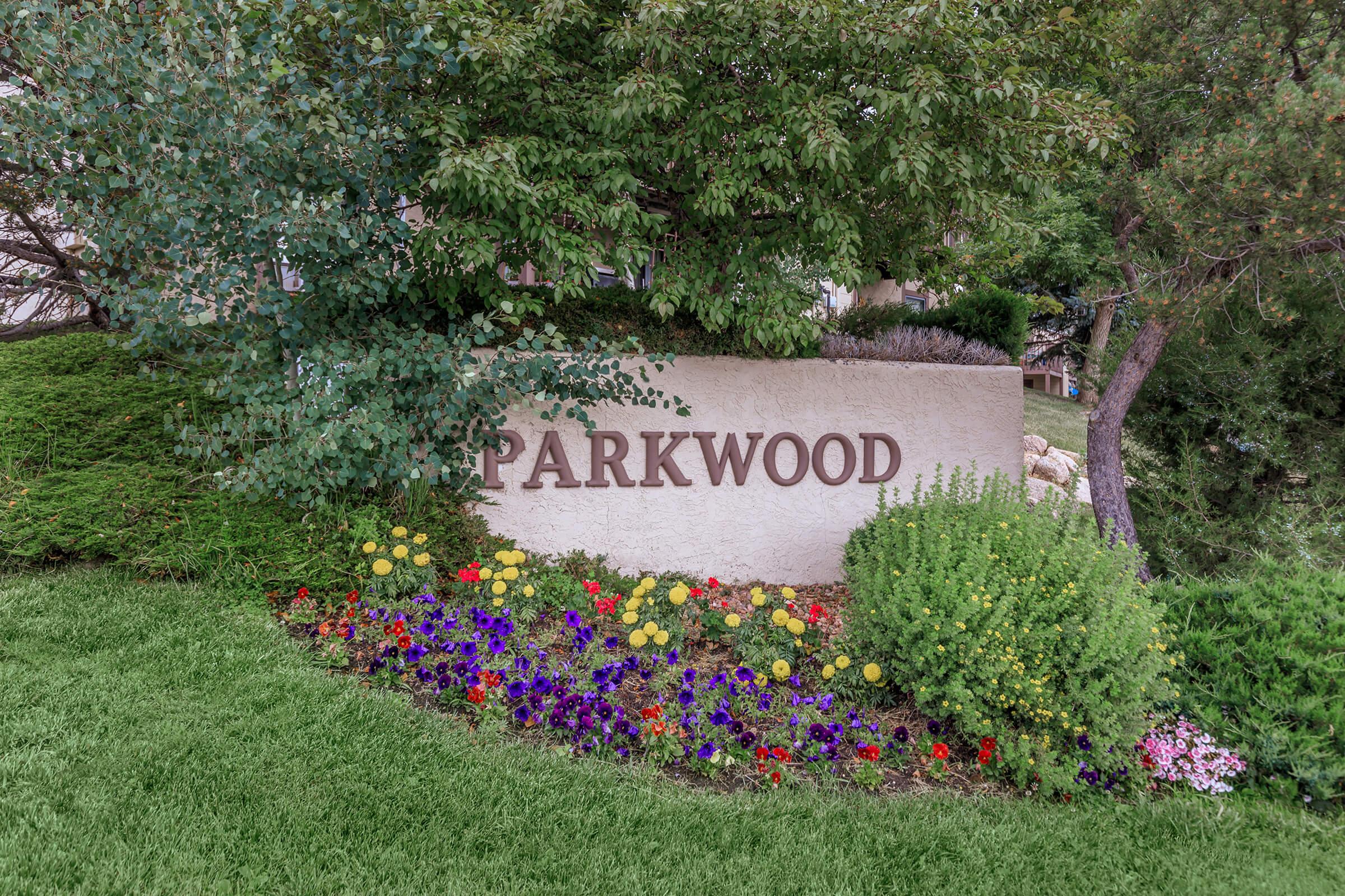 A decorative sign reading "PARKWOOD" is positioned amidst greenery and colorful flower beds. The area features lush plants and vibrant flowers in various colors, creating an inviting atmosphere. Trees are visible in the background, adding to the natural setting.