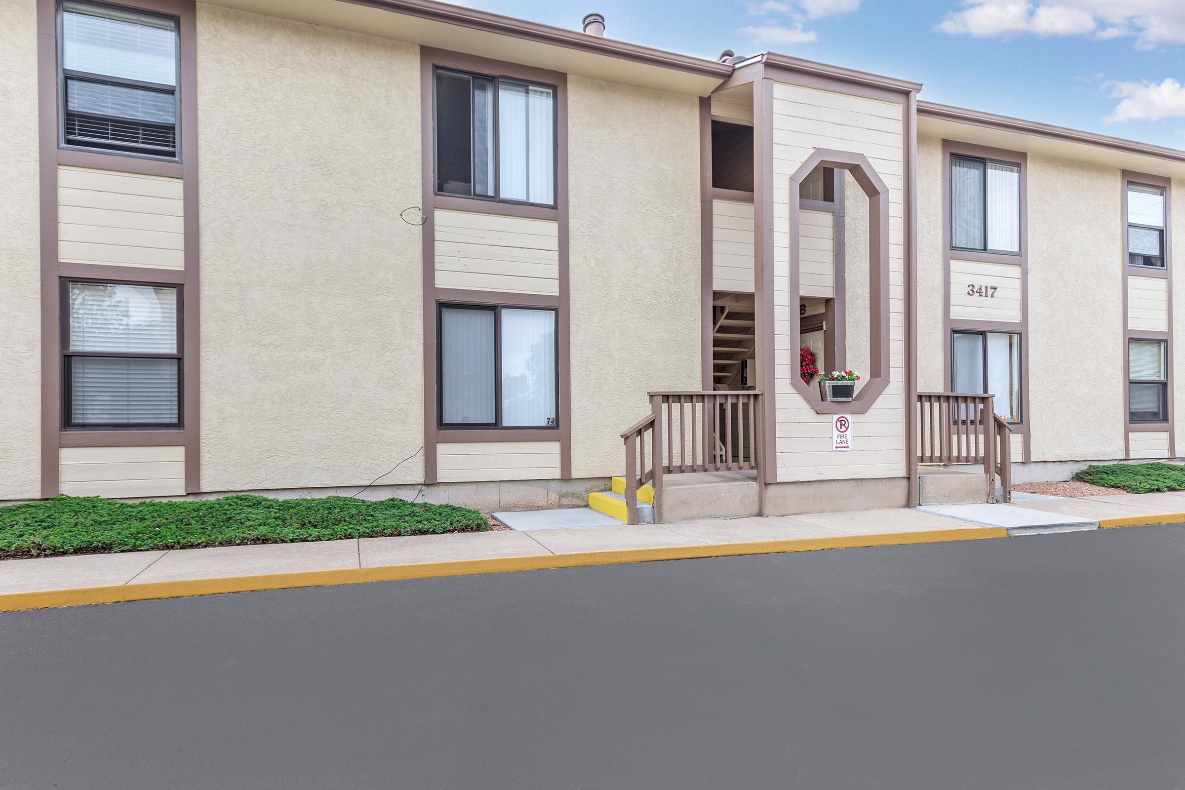 A two-story apartment building with a beige exterior and brown trim. The entrance features a decorative wooden structure and a small set of stairs leading up to the door. There are several windows visible, and the surrounding area has neatly trimmed shrubs and a paved walkway.