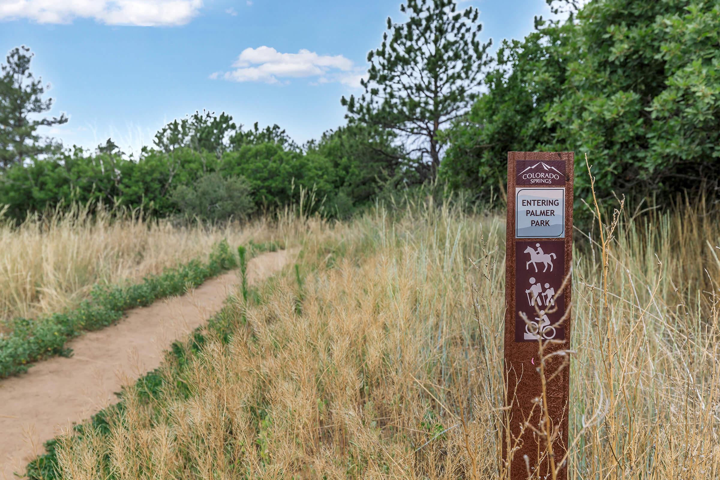 A dirt trail surrounded by tall grass and greenery, leading into Palmer Park. A sign on the left indicates the entrance, featuring icons for walking, horseback riding, and biking. The sky is partly cloudy, adding to the natural setting.