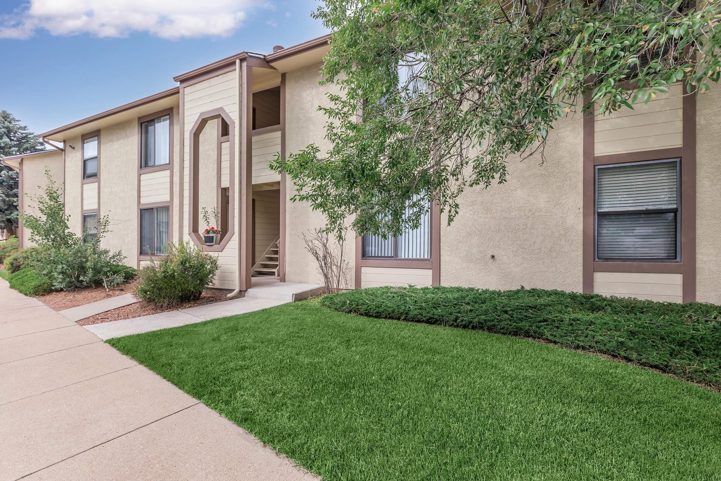 A well-maintained apartment building with a light beige exterior, featuring large windows and a decorative entrance. Lush green grass and shrubs line the walkway, leading to the entrance. The scene is bright and inviting, set against a clear blue sky.
