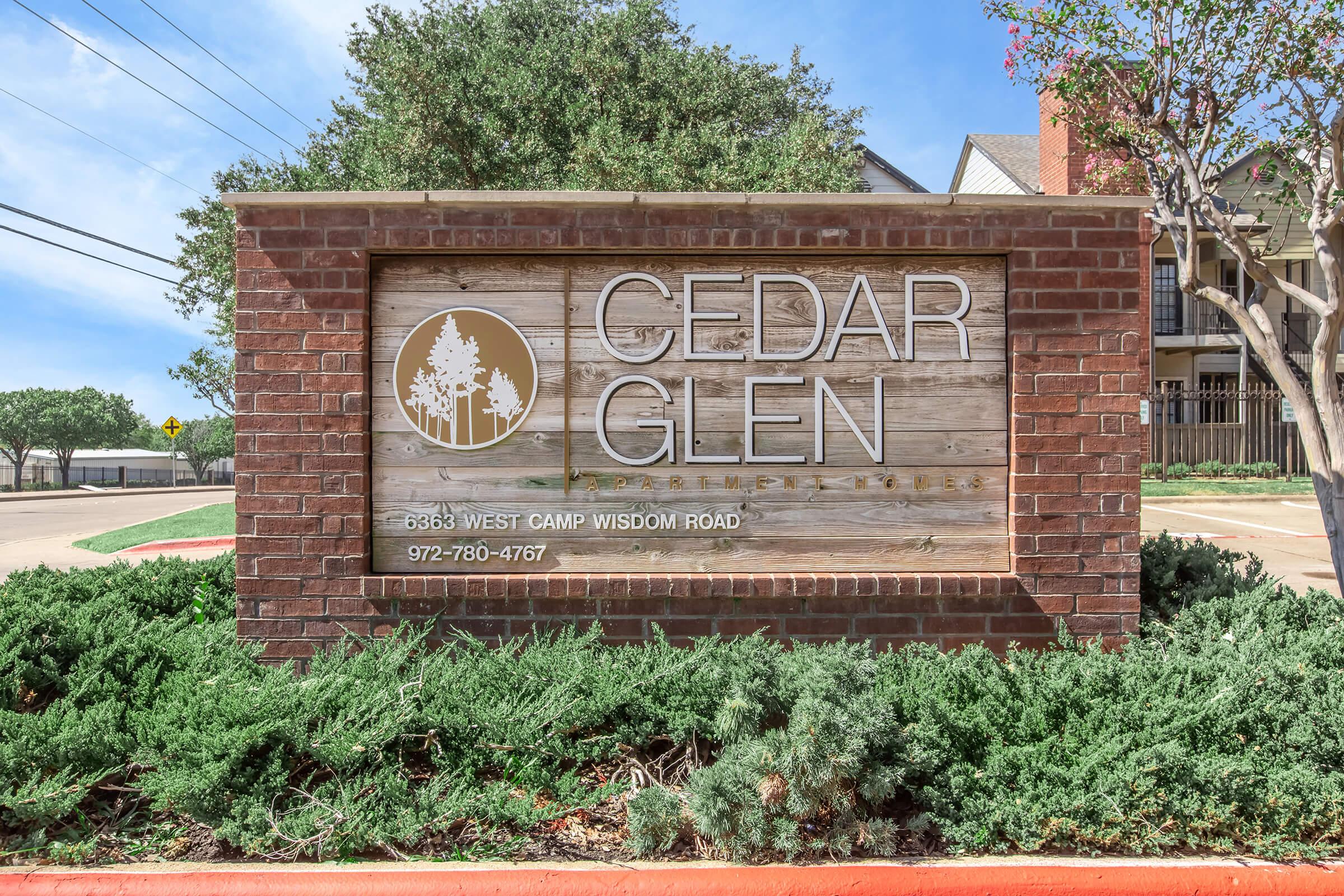 Sign for Cedar Glen Apartments on West Camp Wisdom Road, featuring a wooden design with trees and contact information. Surrounding greenery complements the sign, and the area appears well-maintained with a clear blue sky.
