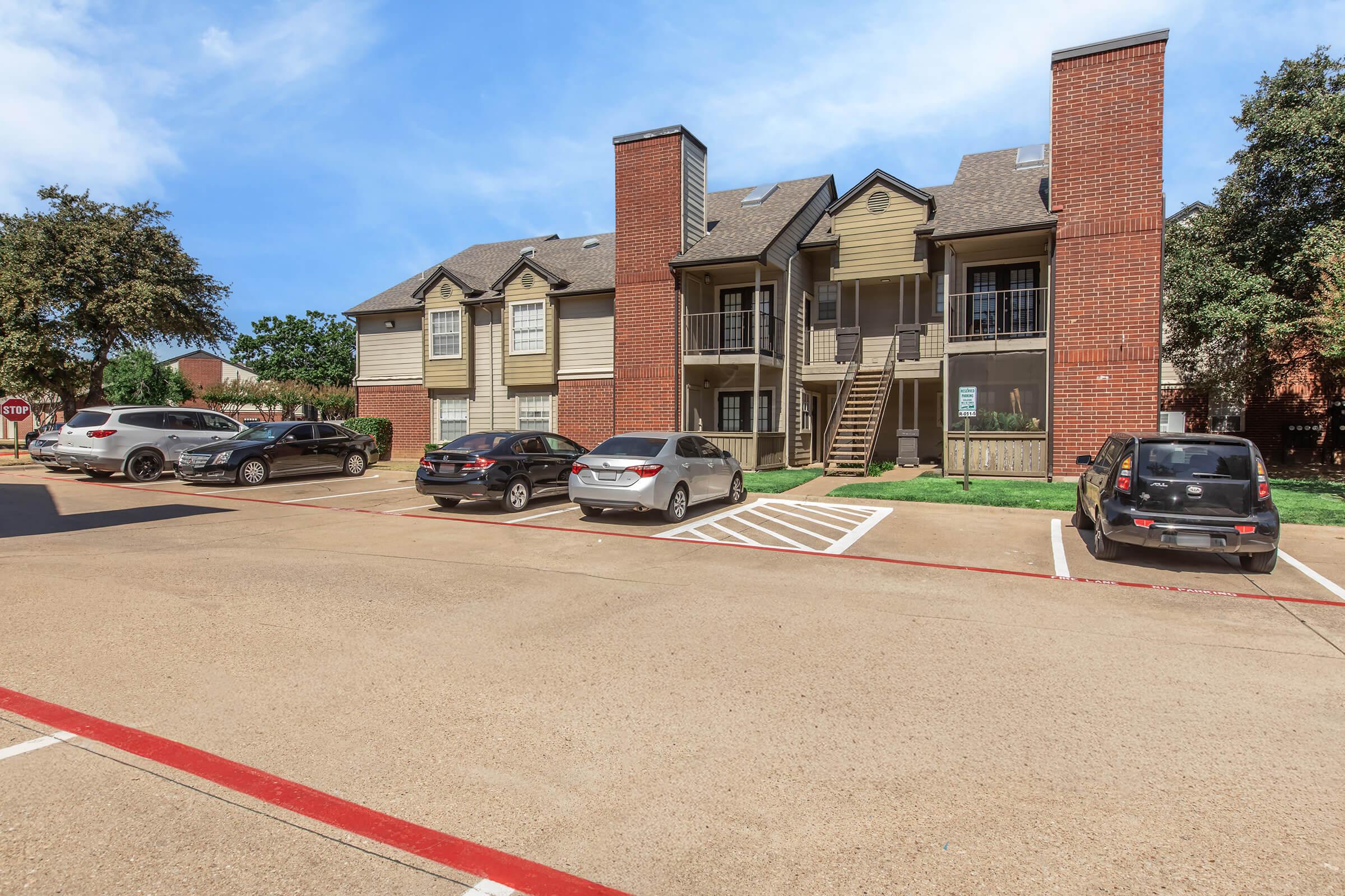 A multi-story apartment complex with a brick facade and wooden balconies, surrounded by a parking lot. Several cars are parked in front, and there are trees and blue sky in the background, creating a pleasant residential atmosphere.