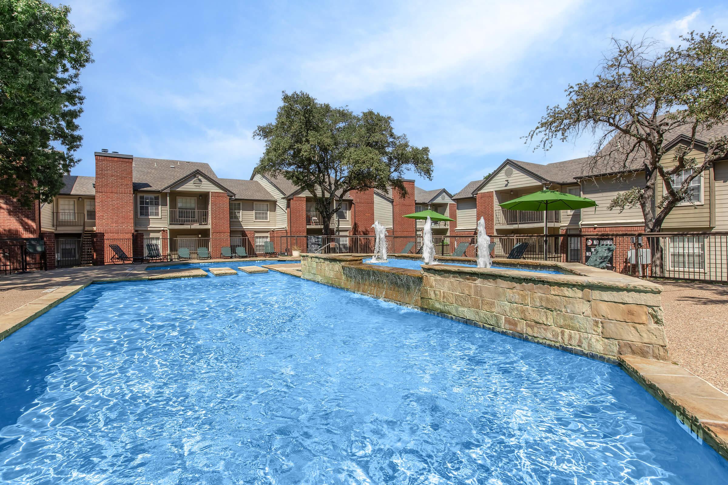 A sparkling blue swimming pool with a stone edge features decorative fountains. Surrounding the pool are green umbrellas and landscaped areas. In the background, there are residential buildings with red brick and beige exteriors, set among trees under a clear blue sky.