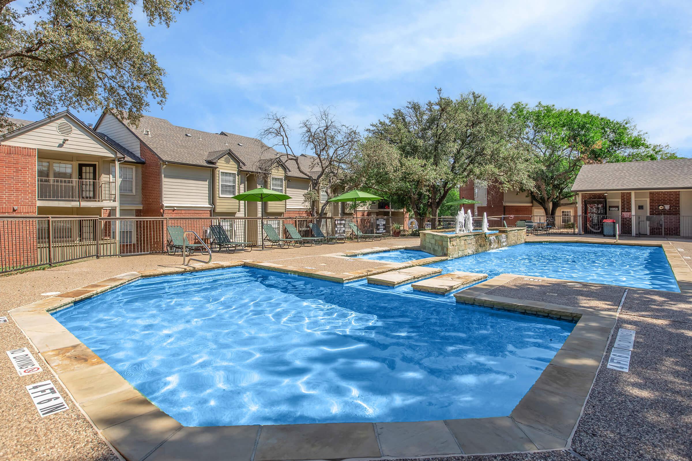 A sunny outdoor pool area featuring two blue swimming pools, surrounded by green trees and seating. There are lounge chairs and umbrellas nearby, with a fountain in one pool. Residential buildings are in the background, providing a relaxing atmosphere.