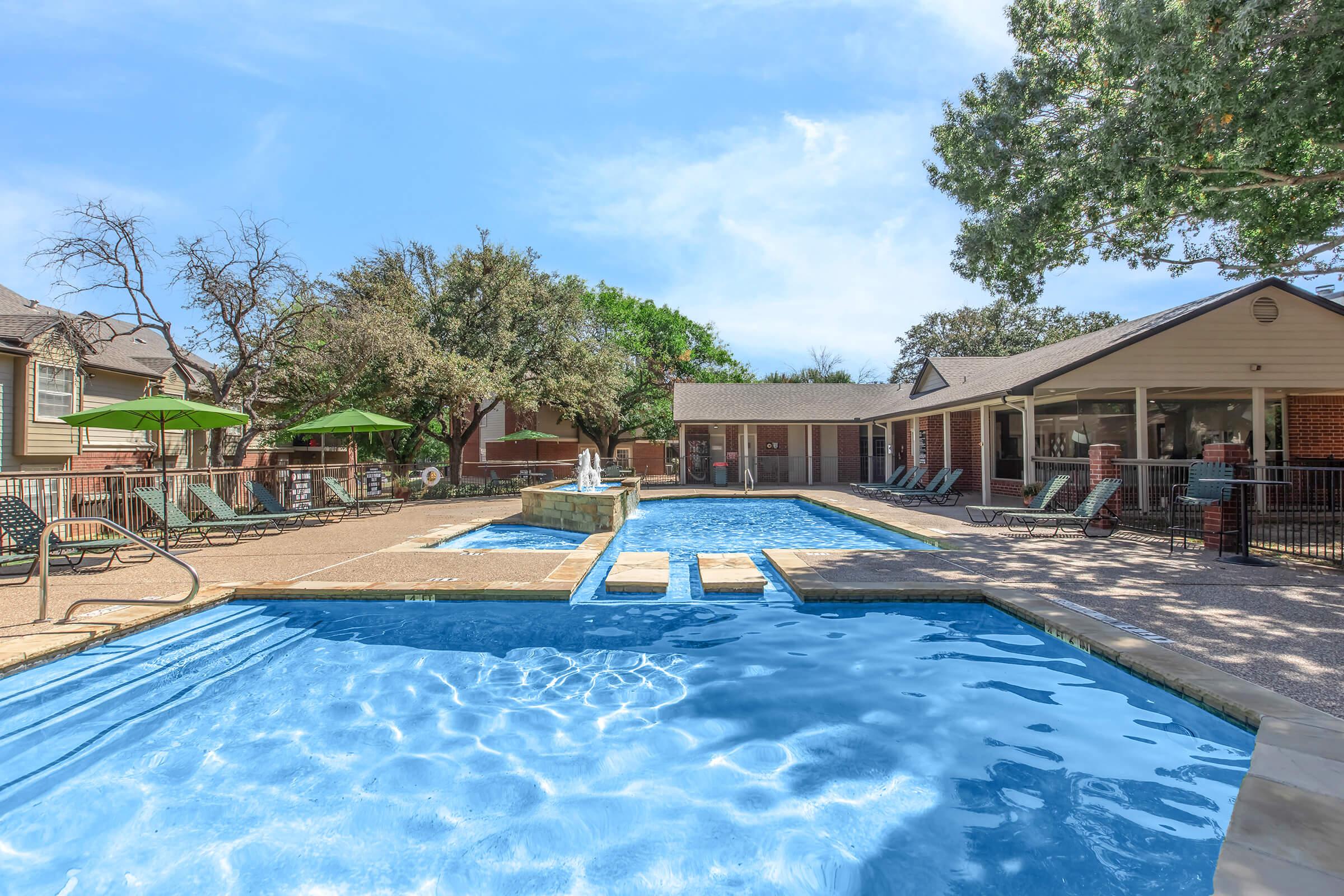 A sunny outdoor swimming pool area featuring two interconnected pools with clear blue water. Surrounding the pools are lounge chairs and green umbrellas. In the background, there are trees and a building with large windows, providing a relaxing atmosphere.