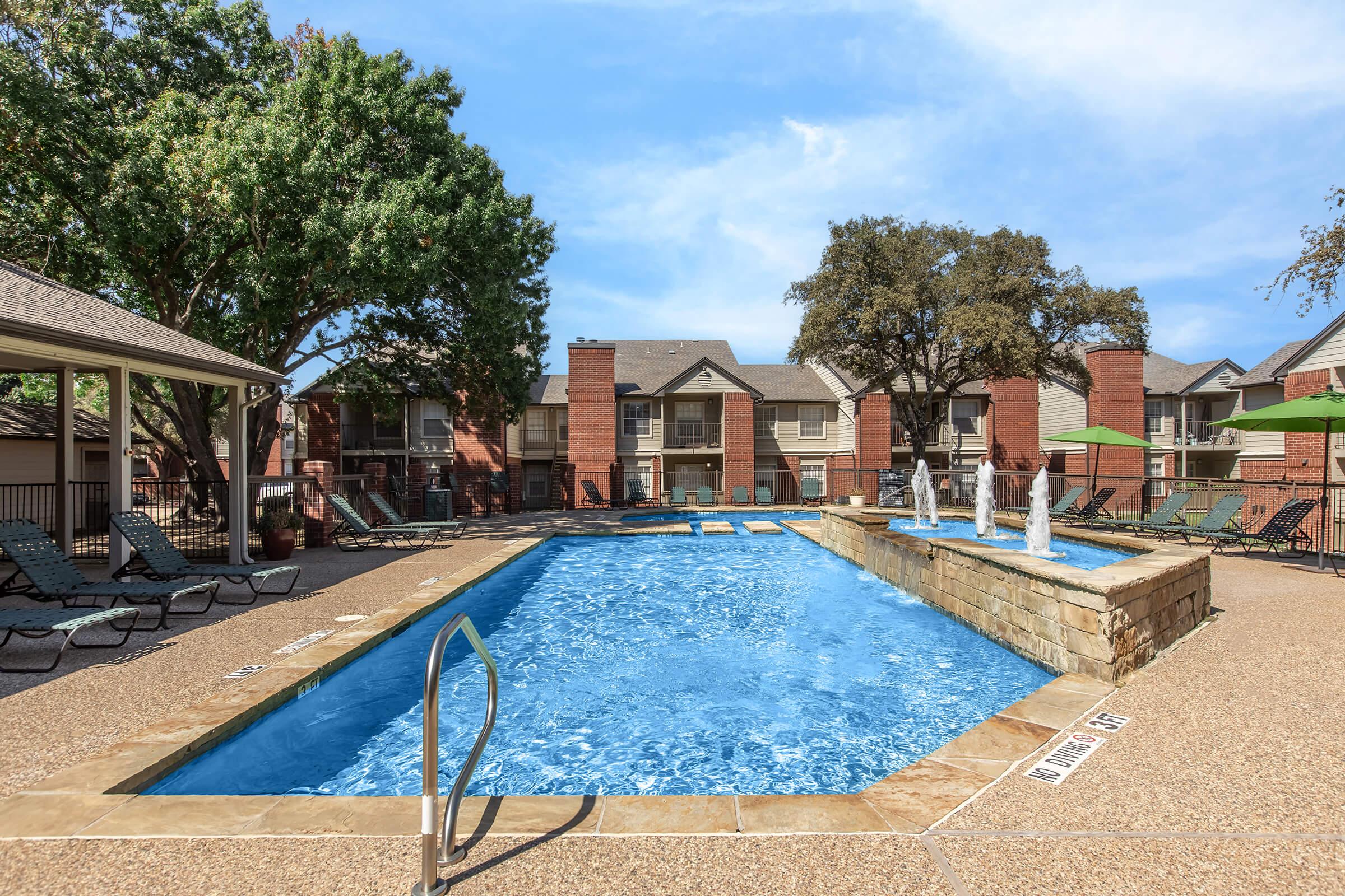 A vibrant swimming pool surrounded by lounge chairs, shaded by large trees. The pool features fountains and has a stone edge. In the background, there are apartment buildings with balconies, and clear blue skies above, creating a relaxing outdoor atmosphere.