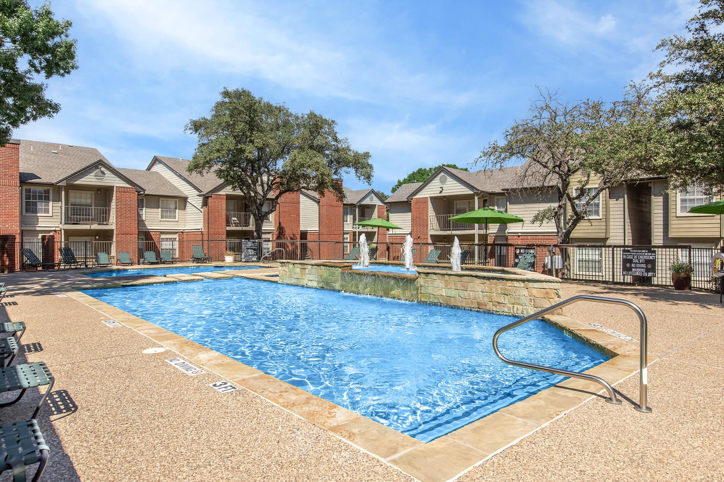 A sunny outdoor swimming pool surrounded by apartment buildings. The pool features a stone fountain and is lined with green umbrellas and lounge chairs. Lush trees provide shade, and the clear blue sky enhances the inviting atmosphere.