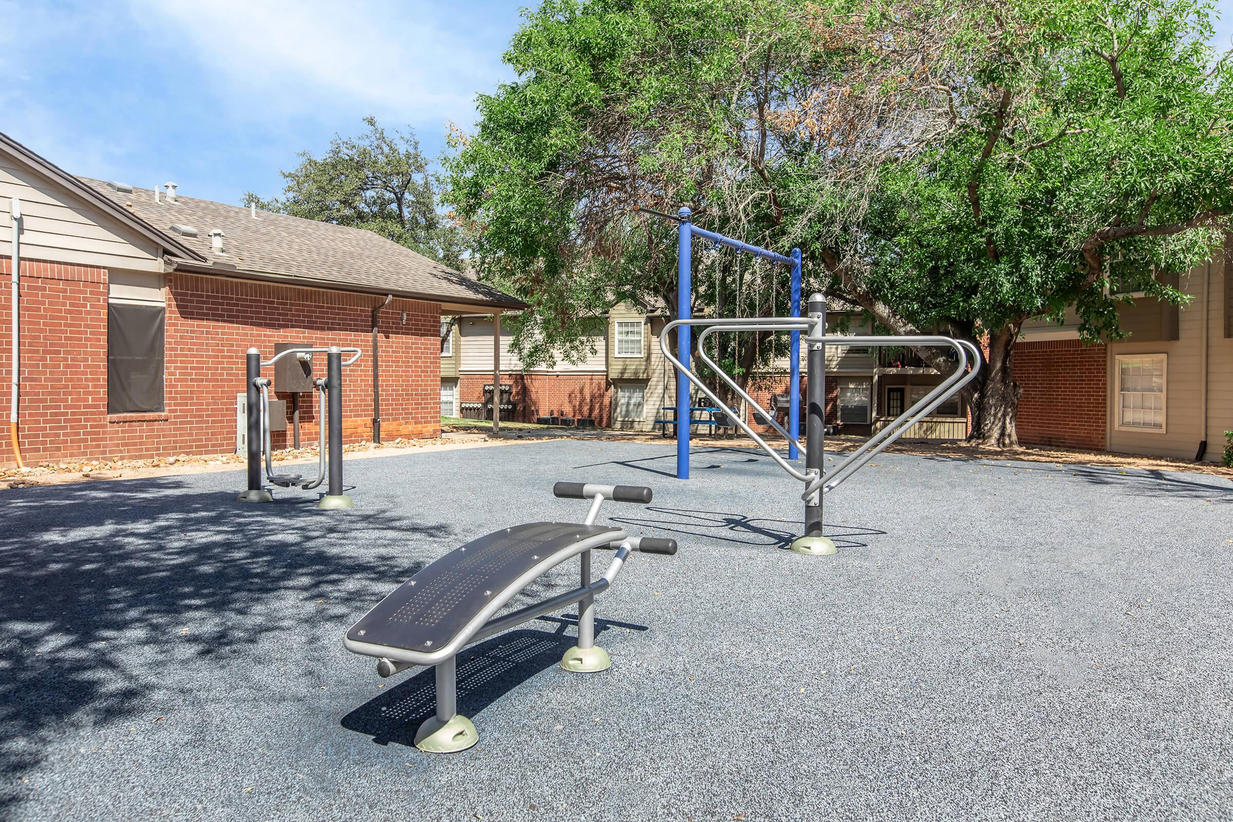 An outdoor fitness area featuring exercise equipment, including a bench for sit-ups and a multi-use pull-up station, situated on a soft, rubberized surface surrounded by trees and residential buildings. Bright blue sky enhances the open, inviting atmosphere.