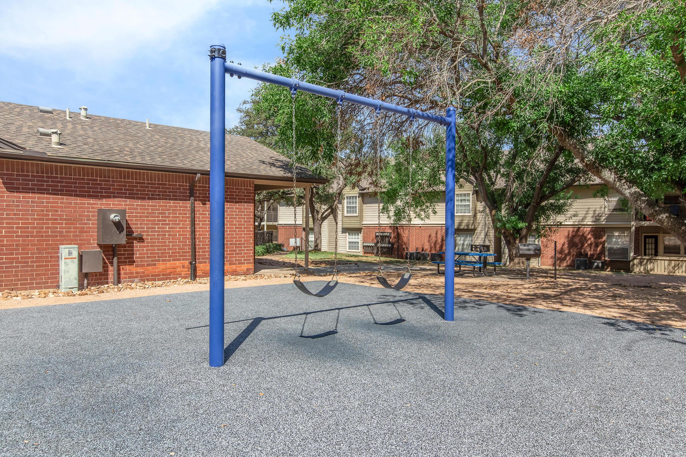 A blue swing set with two empty swings located in a play area surrounded by trees and residential buildings. The ground is covered with a rubber surface, and there is a parking area visible nearby.