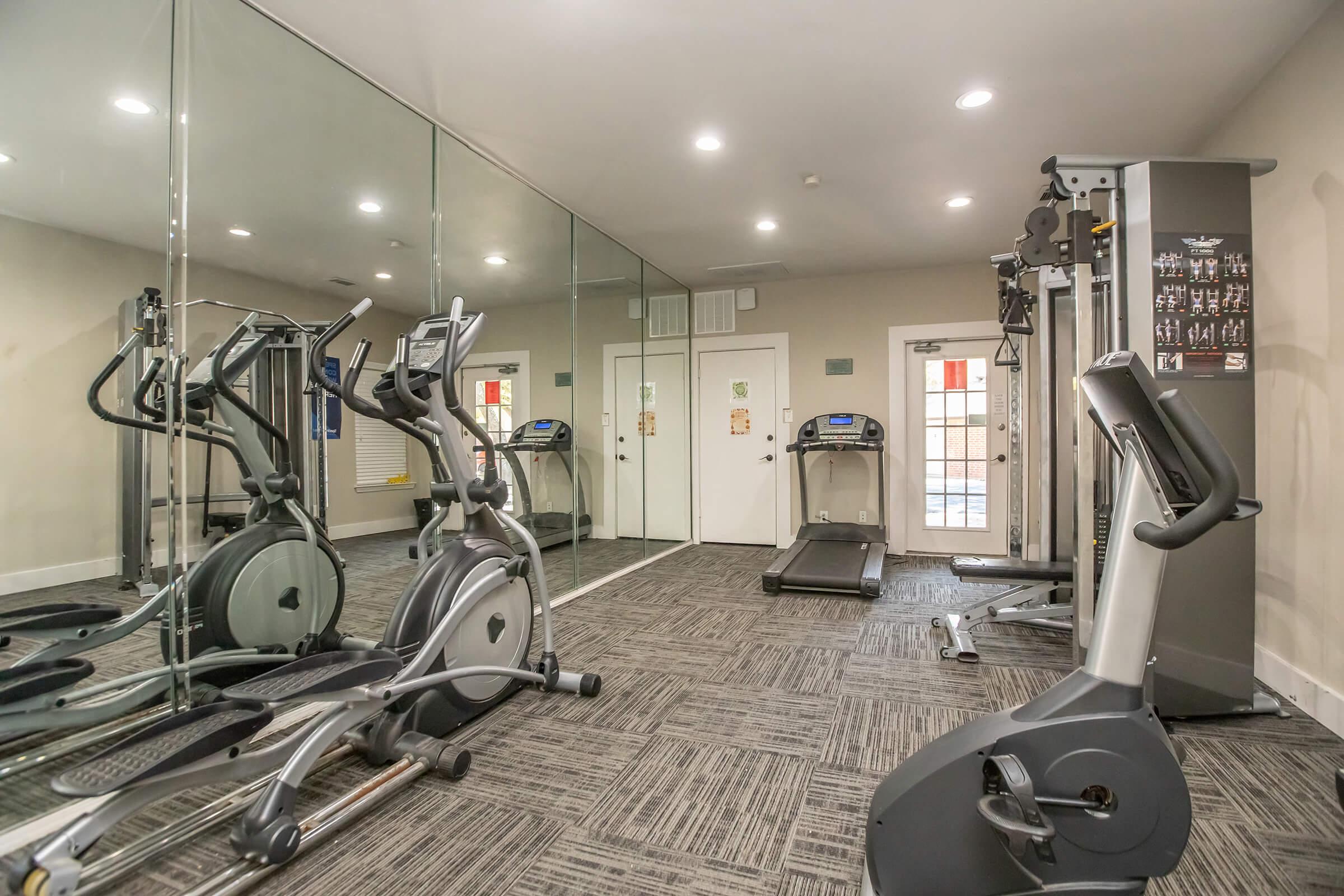 A modern fitness room featuring exercise equipment including an elliptical machine, a treadmill, and a multi-station gym machine. The space is well-lit, with mirrors covering one wall and a clean, carpeted floor. There are doors leading outside visible through a glass panel.