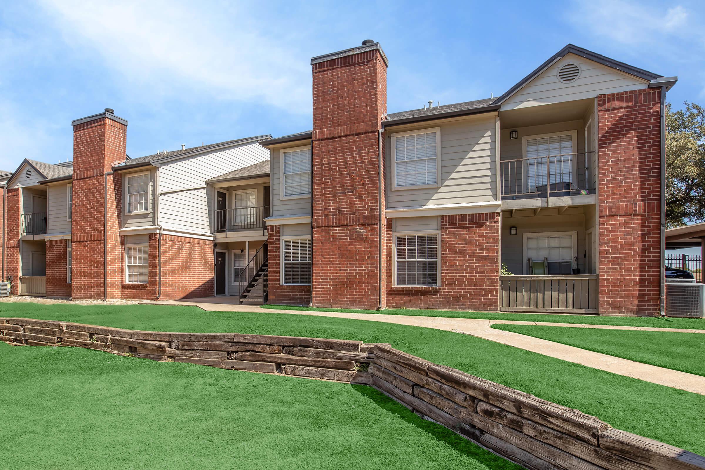 A two-story apartment building featuring red brick and beige siding, with balconies and large windows. The building is set in a well-maintained grassy area with a wooden fence in the foreground. The sky is bright and clear, indicating a sunny day.