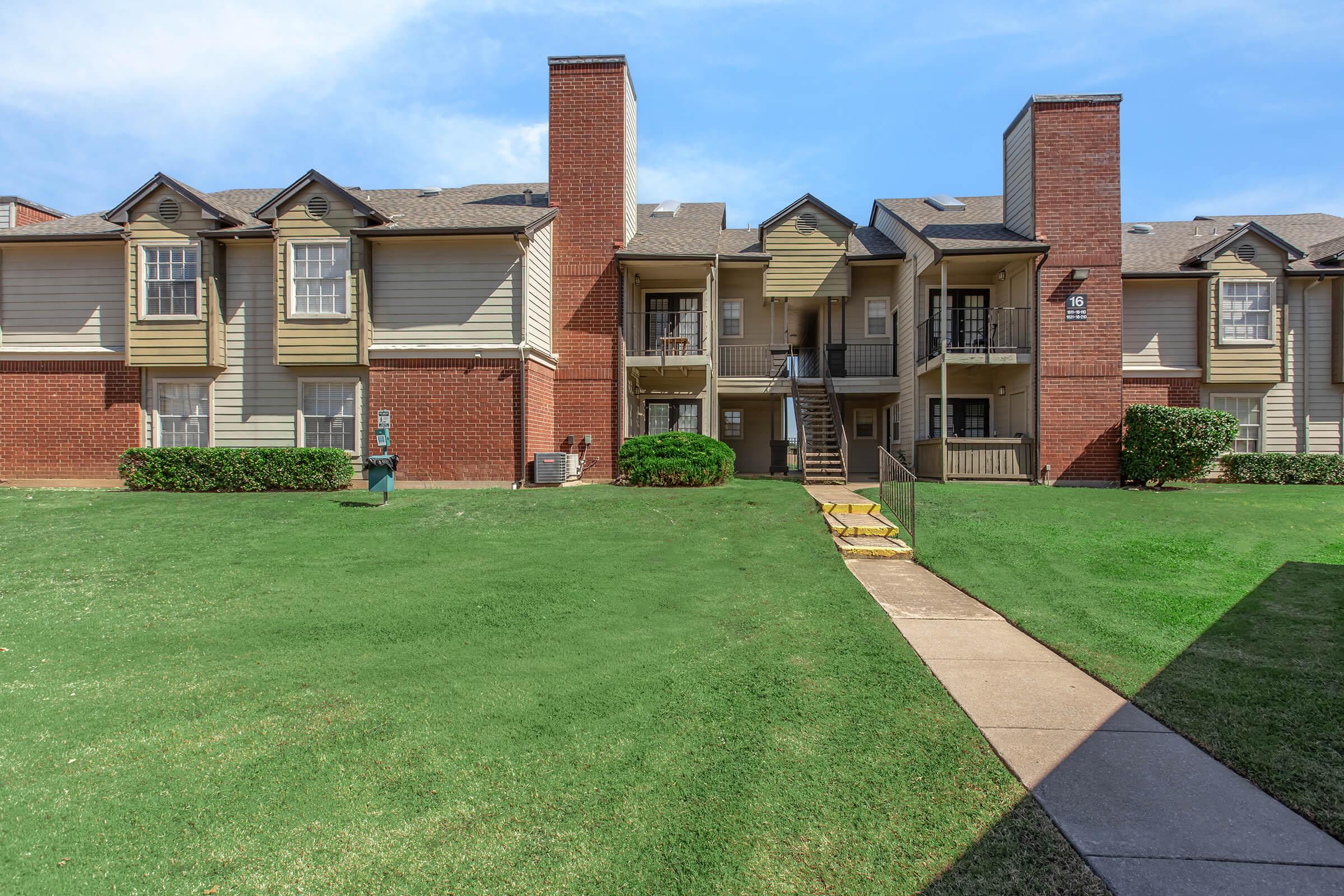 A view of a residential apartment complex featuring two-story buildings with a mix of brick and wooden siding. The landscaping includes well-maintained green lawns and shrubs. There are staircases leading to upper-level apartments, and a clear blue sky is visible in the background.