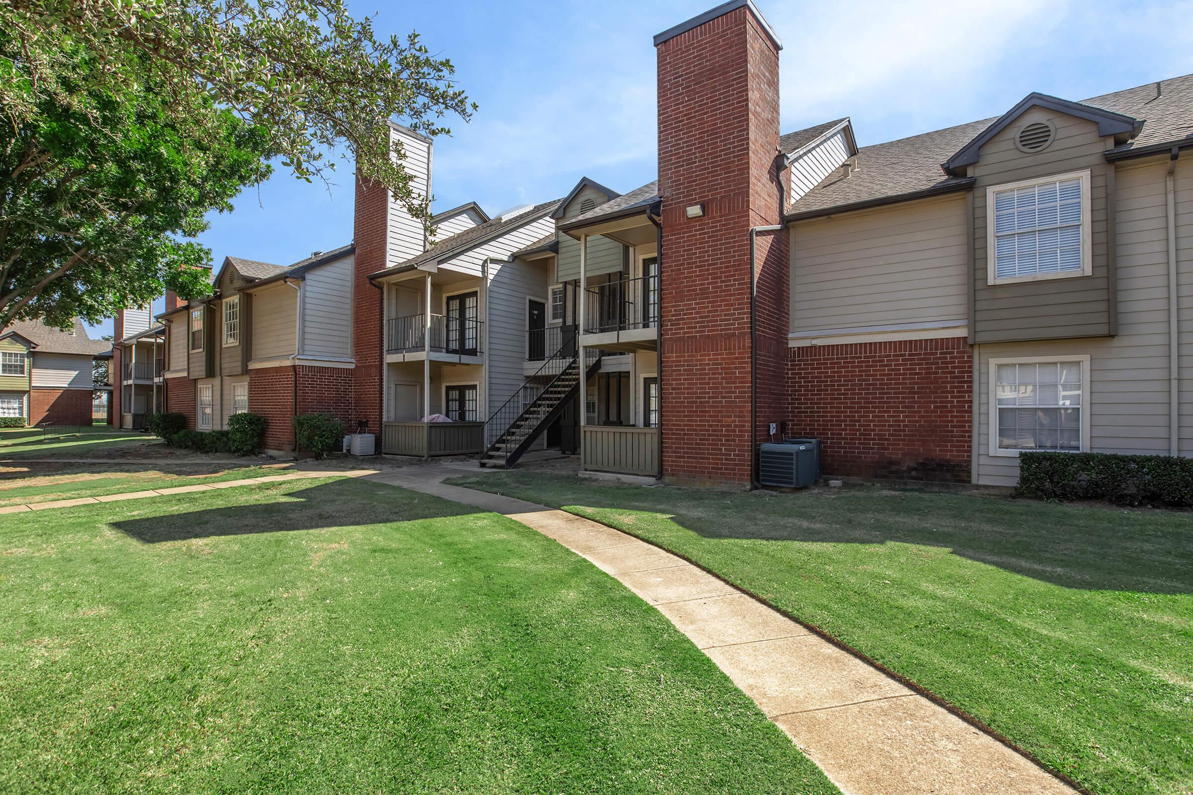 A residential area featuring multiple apartment buildings with brick and wooden facades. The buildings have balconies and are surrounded by well-maintained green lawns. A concrete pathway winds through the landscape, leading to the entrances of the apartments. Trees provide shade in the vicinity.