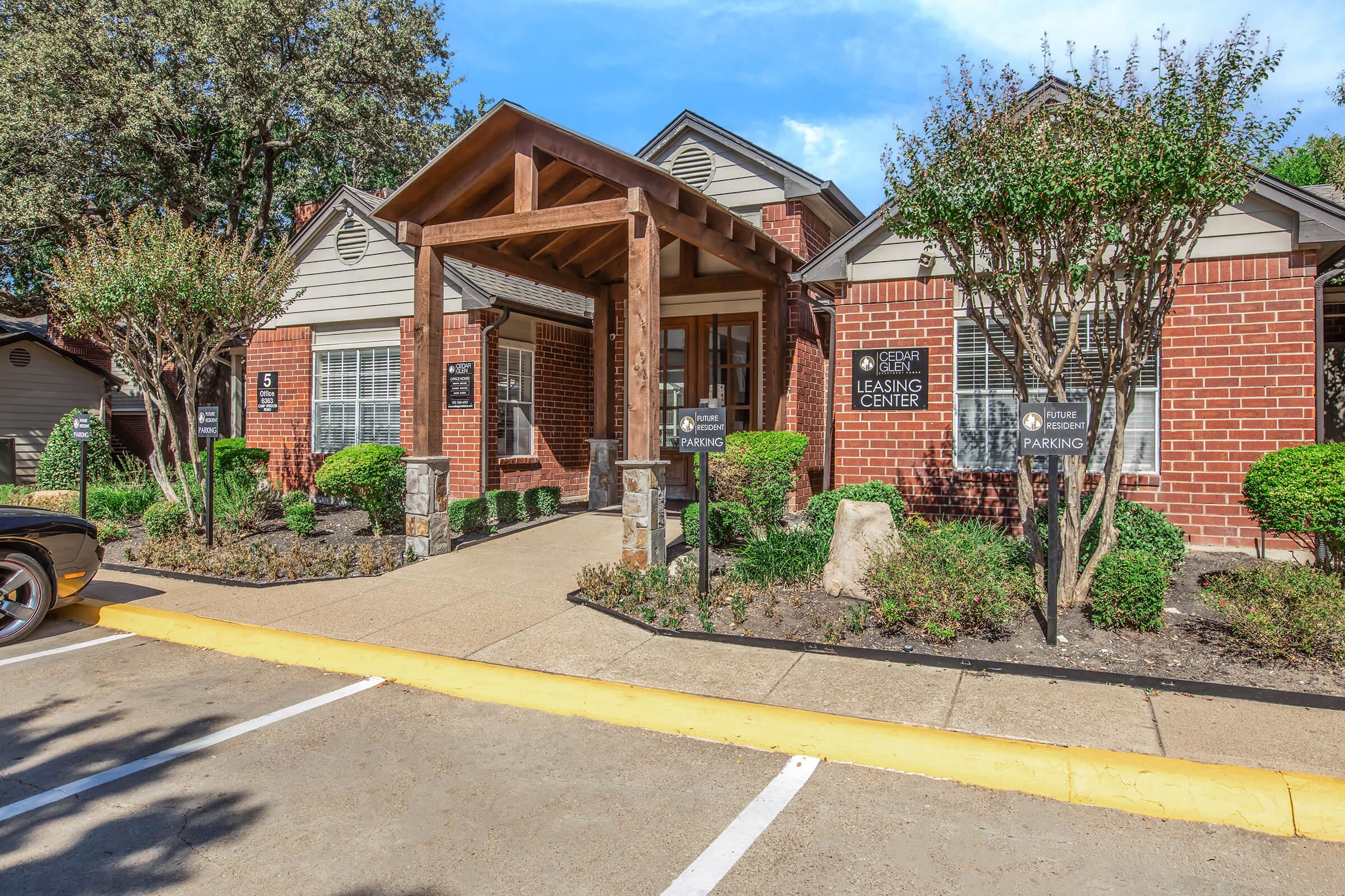 A brick leasing center building with a wooden overhang entrance, surrounded by neatly trimmed bushes and small trees. A paved path leads to the entrance, with parking signs visible in the foreground. The sky is clear and blue, creating a welcoming atmosphere.