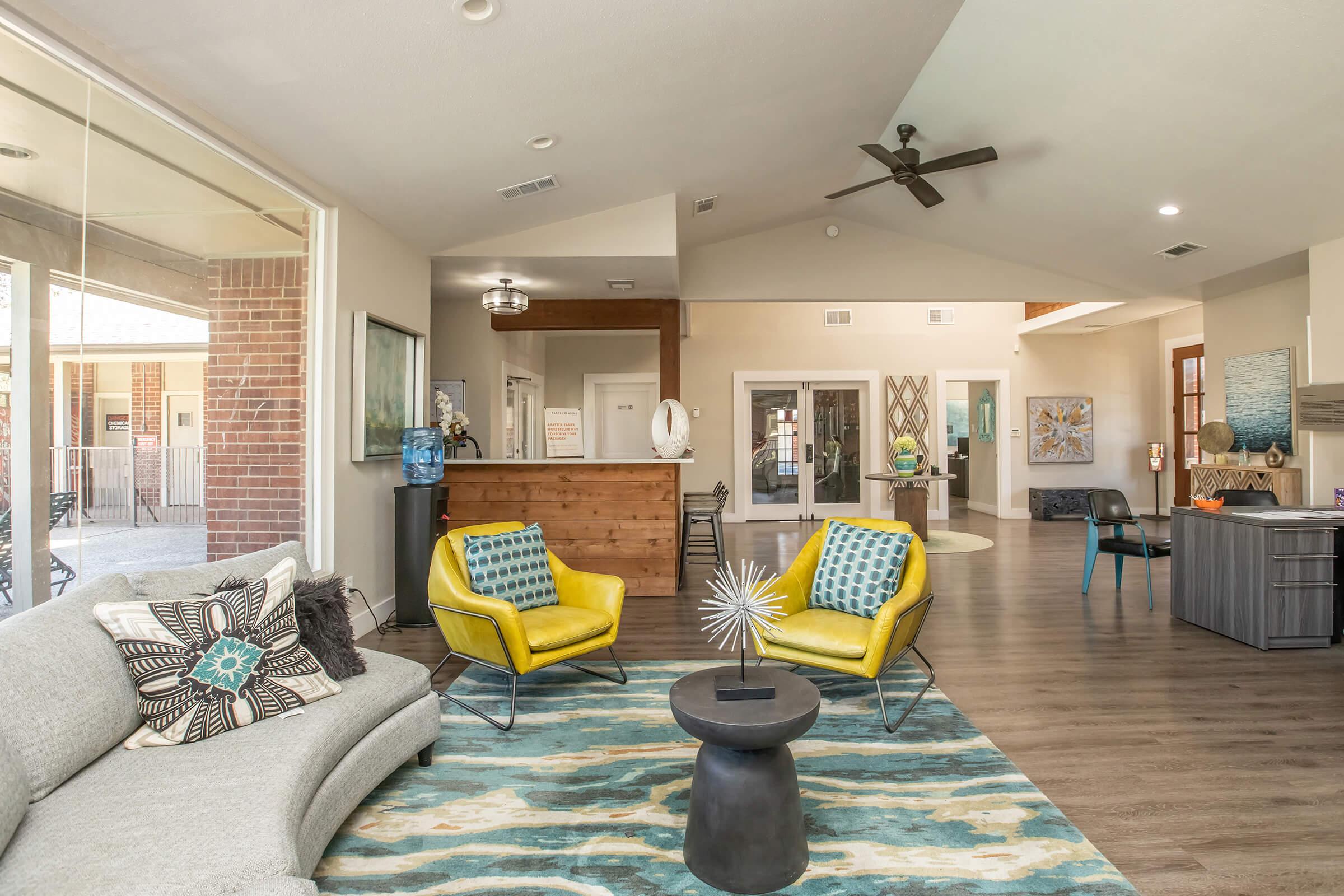 A modern indoor lounge area featuring a light gray sofa, two bright yellow armchairs, and a dark circular coffee table. The space has wood flooring, large windows letting in natural light, decorative artwork on the walls, and a ceiling fan. A welcoming atmosphere with contemporary design elements.