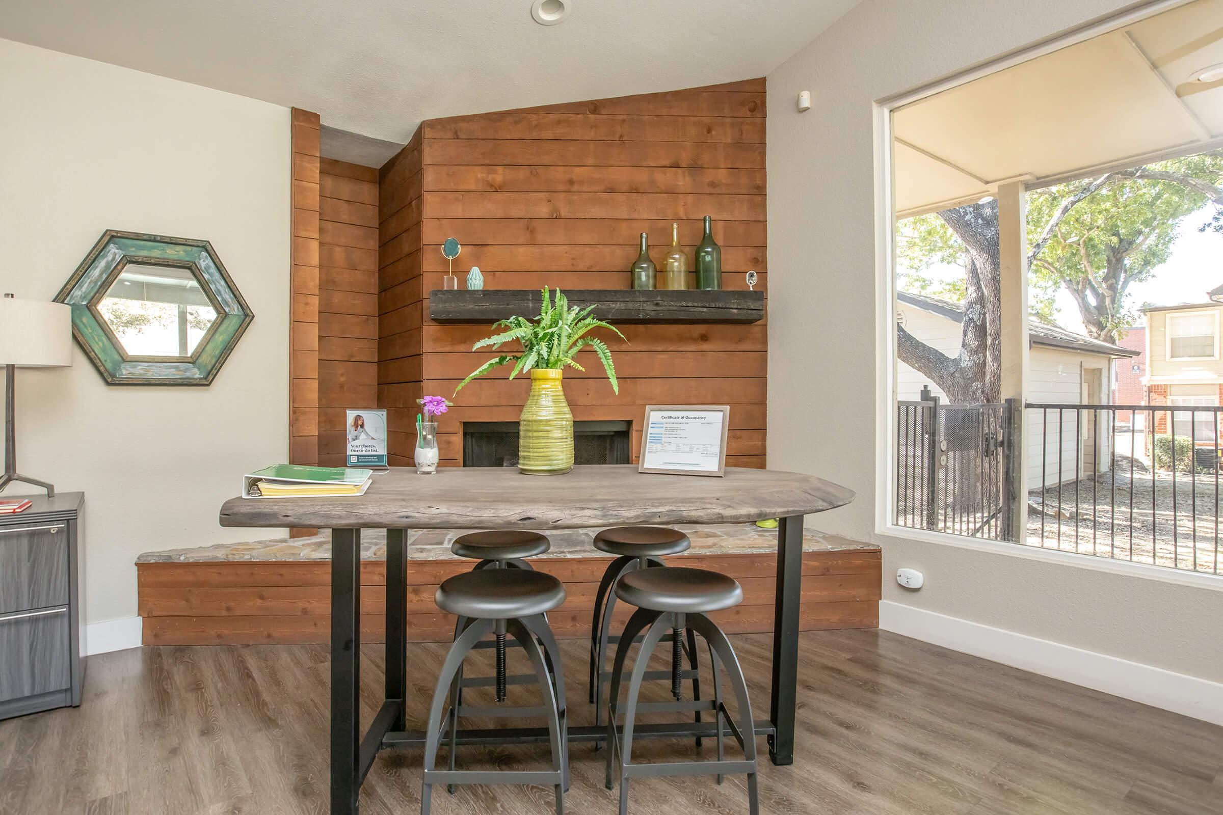 A cozy indoor seating area featuring a wooden table with three black stools. There's a plant centerpiece on the table, a decorative mirror on the wall, and a shelf with glass bottles. Natural light comes through a nearby window, illuminating rustic wood paneling in the background.